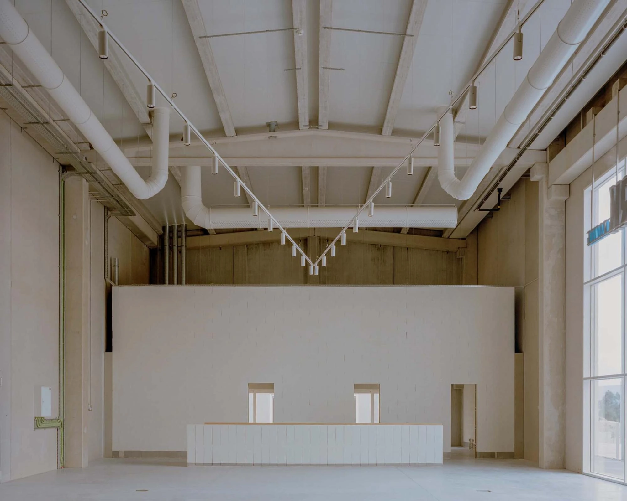 Corcet Headquarters by Nuno M. Sousa, photographed by Federico Farinatti, Francisco Ascensão — White ductwork branches across a pitched ceiling above a long counter, two small windows punched into the pale mezzanine wall behind it.