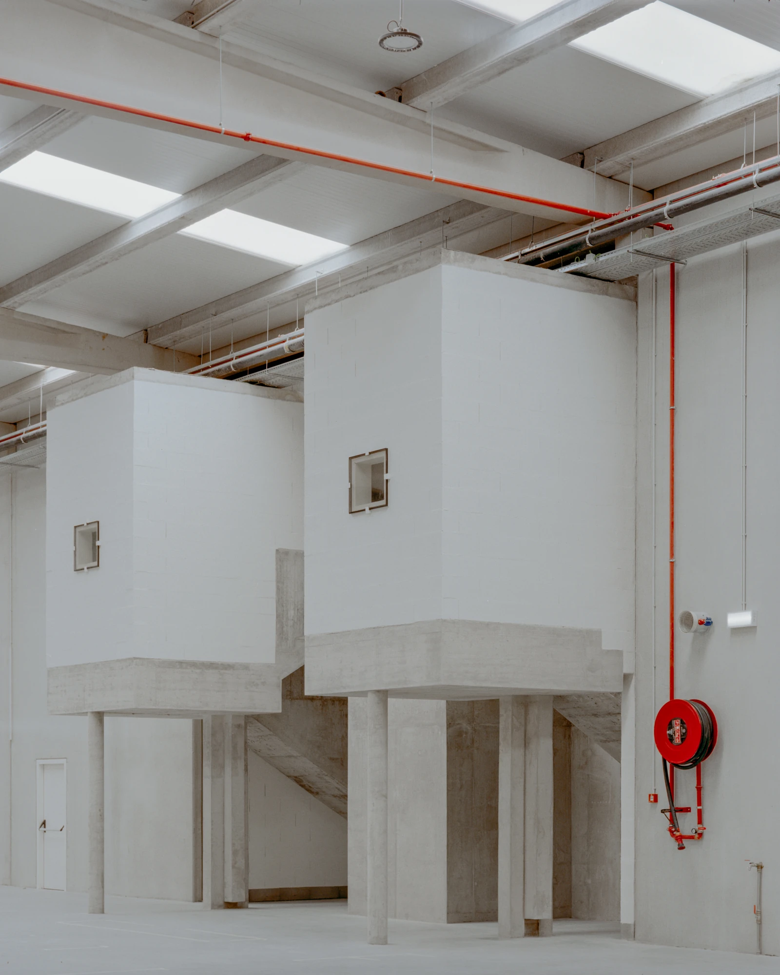 Corcet Headquarters by Nuno M. Sousa, photographed by Federico Farinatti, Francisco Ascensão — Two pale boxes ride on concrete supports above the hall floor, each pierced by a small square window, a red hose reel mounted on the wall alongside.