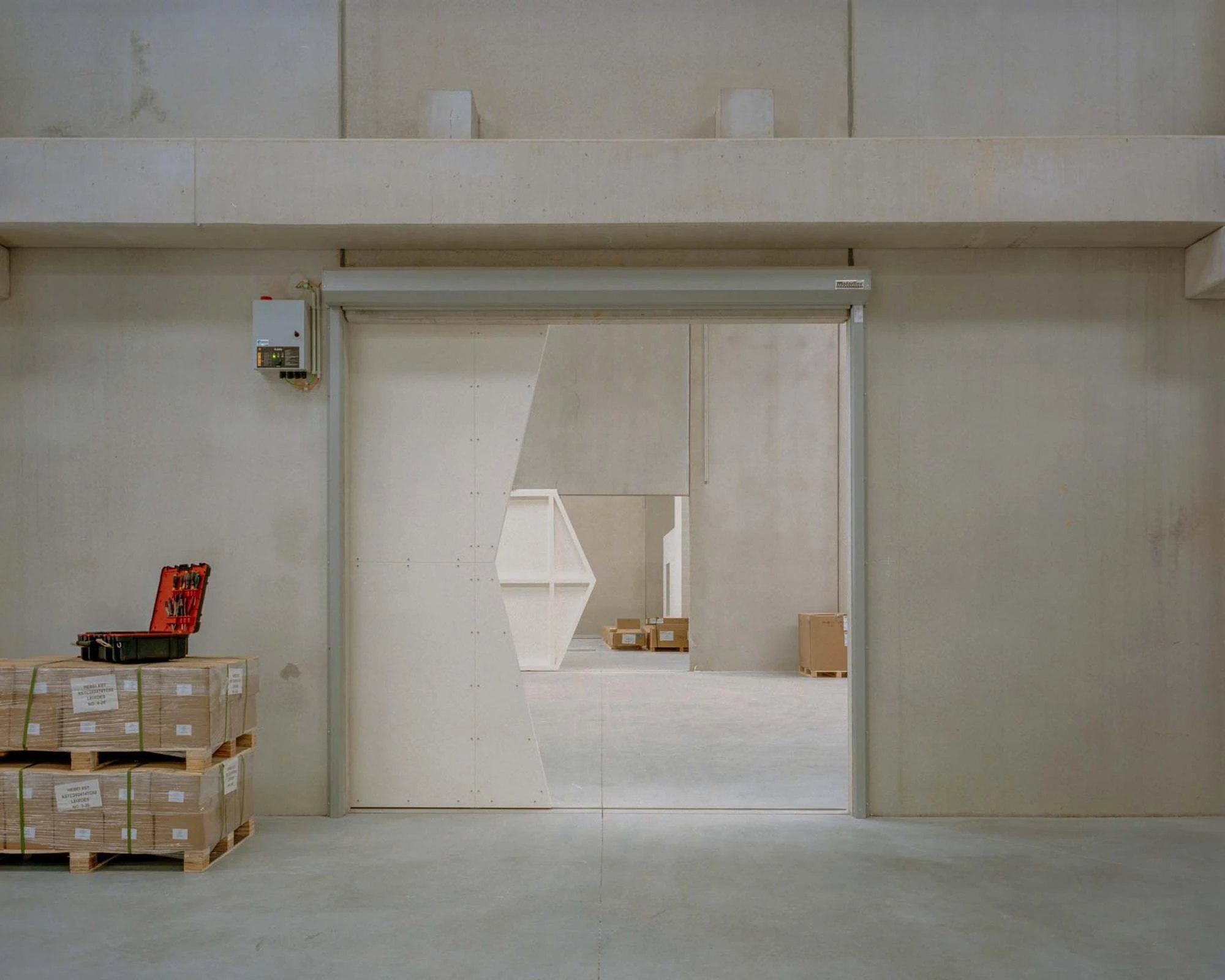 Corcet Headquarters by Nuno M. Sousa, photographed by Federico Farinatti, Francisco Ascensão — A broad sliding door stands open onto a storage bay, a white triangular frame and palletised crates visible in the room beyond.