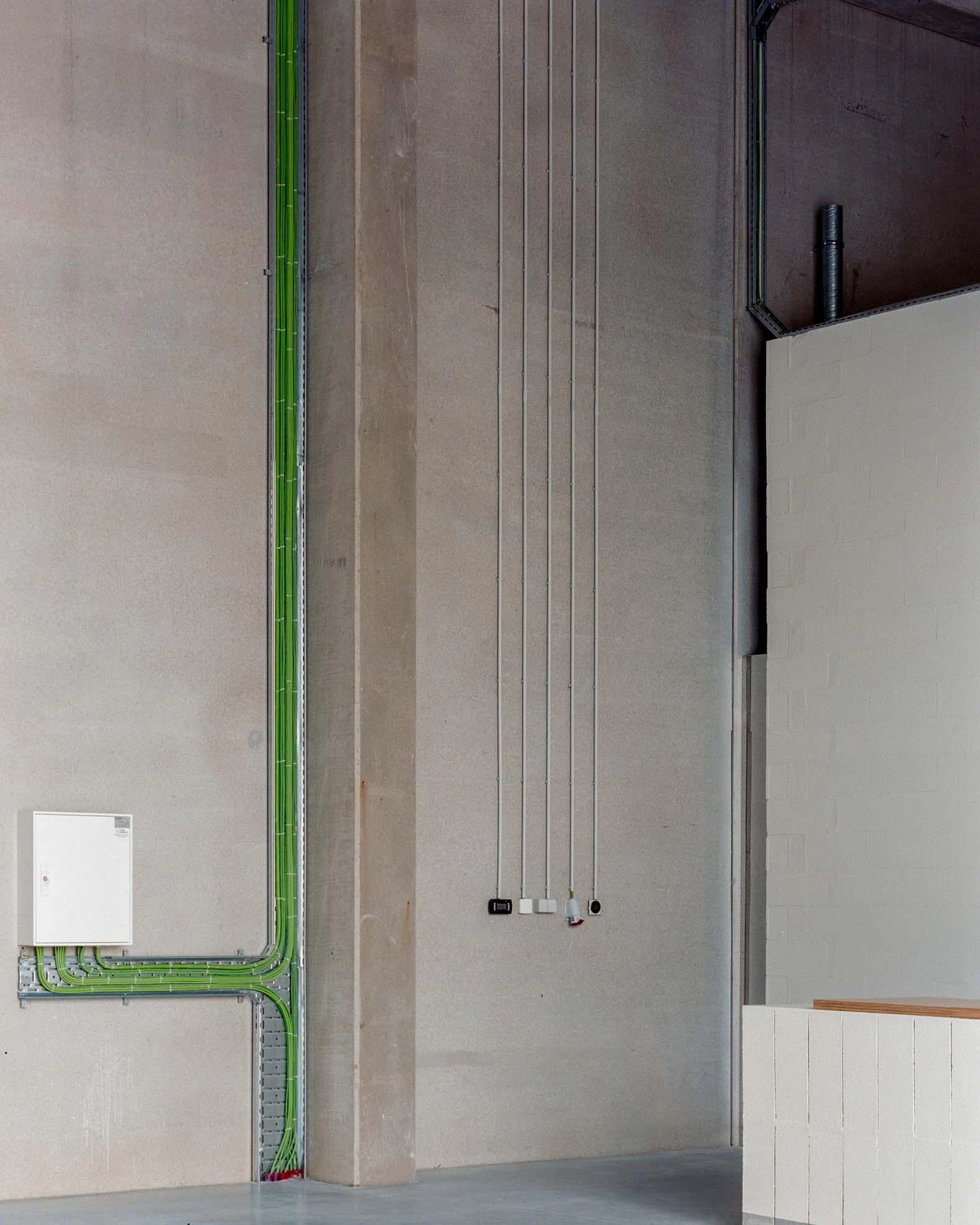 Corcet Headquarters by Nuno M. Sousa, photographed by Federico Farinatti, Francisco Ascensão — Green cable trays climb a concrete pillar and fan out along the floor, loose wiring hanging to a row of sockets at waist height.