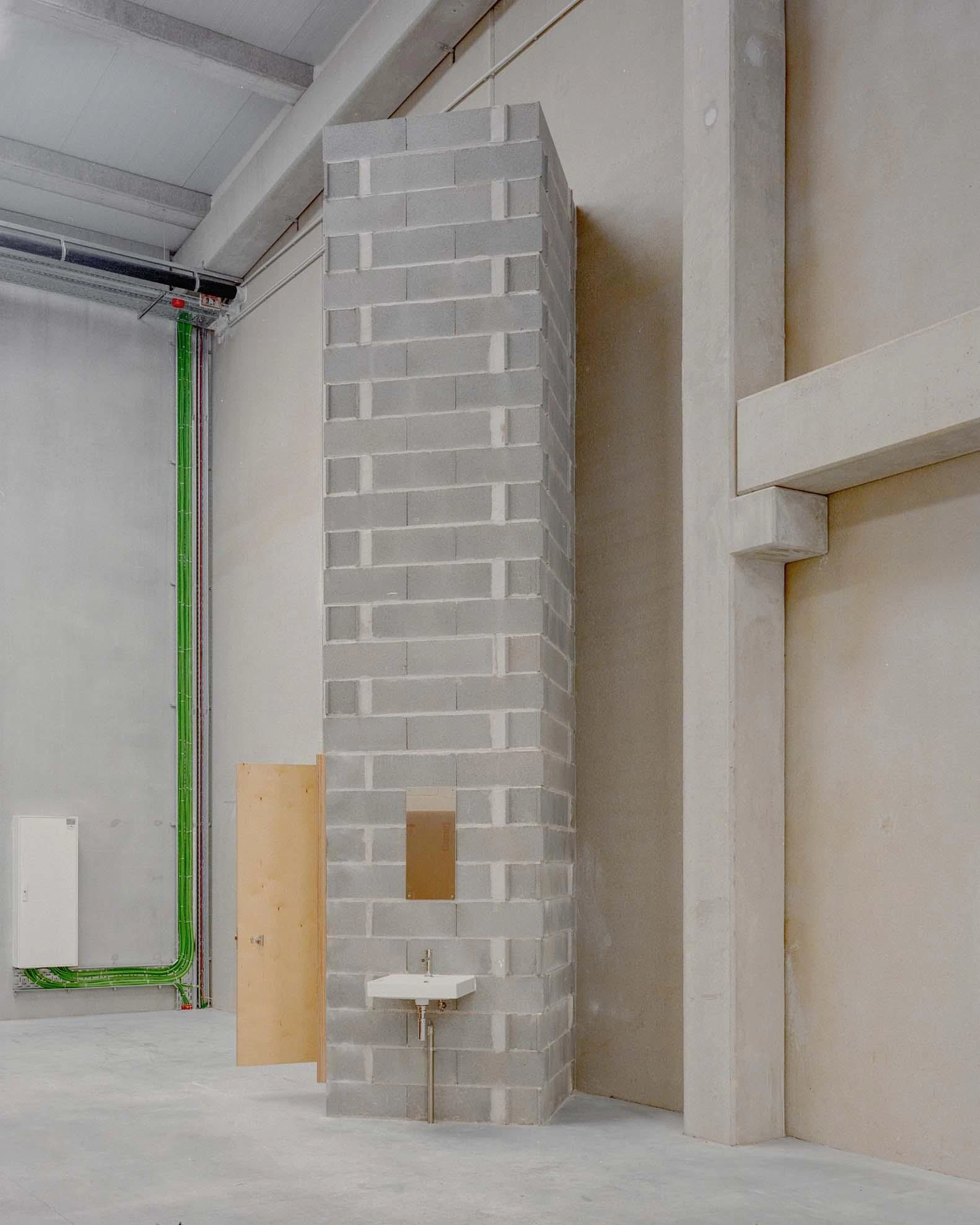 Corcet Headquarters by Nuno M. Sousa, photographed by Federico Farinatti, Francisco Ascensão — A tall stack of pale blocks meets the ceiling beam, a small wall-hung basin and a leaning board set against its base.