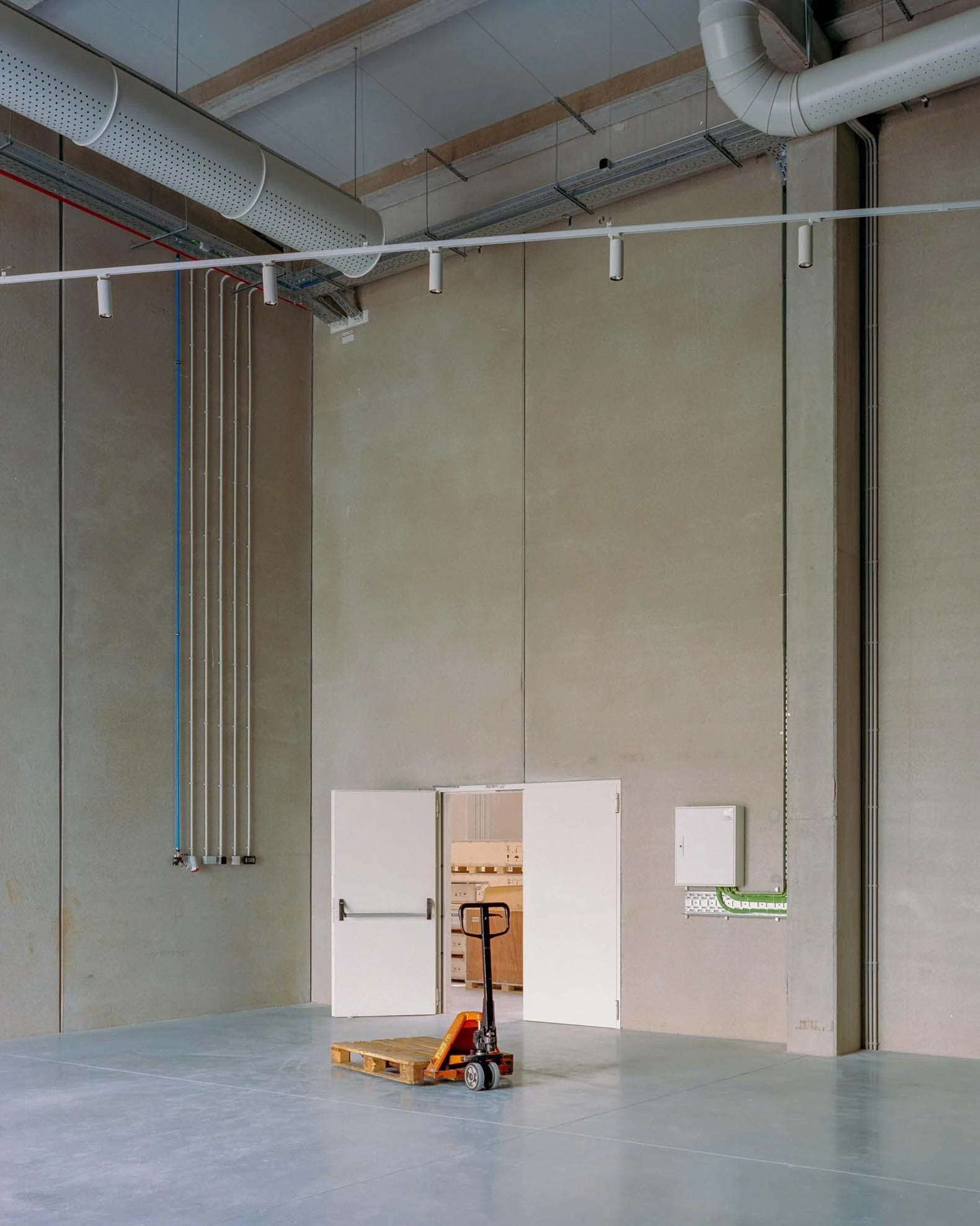 Corcet Headquarters by Nuno M. Sousa, photographed by Federico Farinatti, Francisco Ascensão — A pallet truck waits on the concrete floor before a pair of fire doors, tall bare walls rising to ducts and conduit overhead.