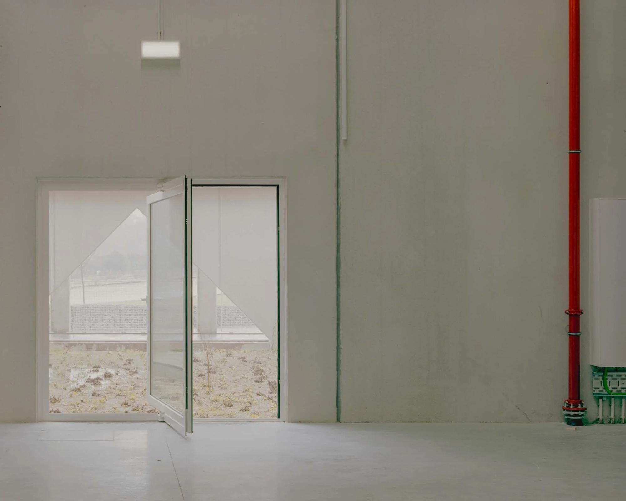 Corcet Headquarters by Nuno M. Sousa, photographed by Federico Farinatti, Francisco Ascensão — A casement swings open to a gravelled courtyard beyond, a red standpipe running floor to ceiling on the pale wall at right.