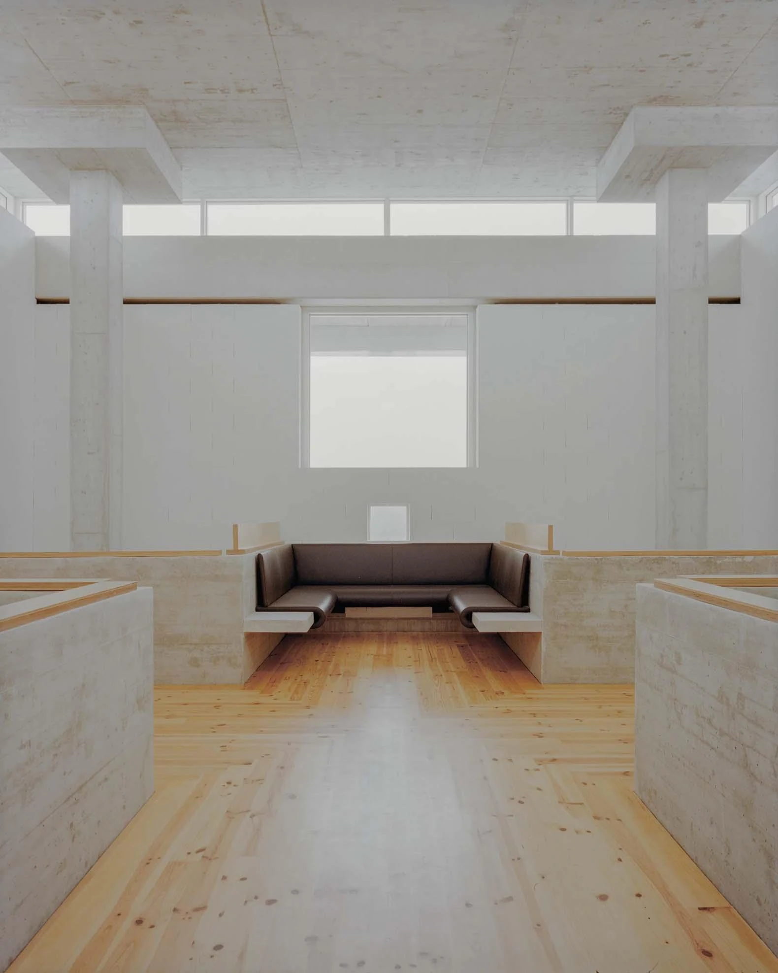 Corcet Headquarters by Nuno M. Sousa, photographed by Federico Farinatti, Francisco Ascensão — A built-in bench nook sits below a bright square window between two low concrete walls, a pale plank floor running toward it under a band of clerestory light.