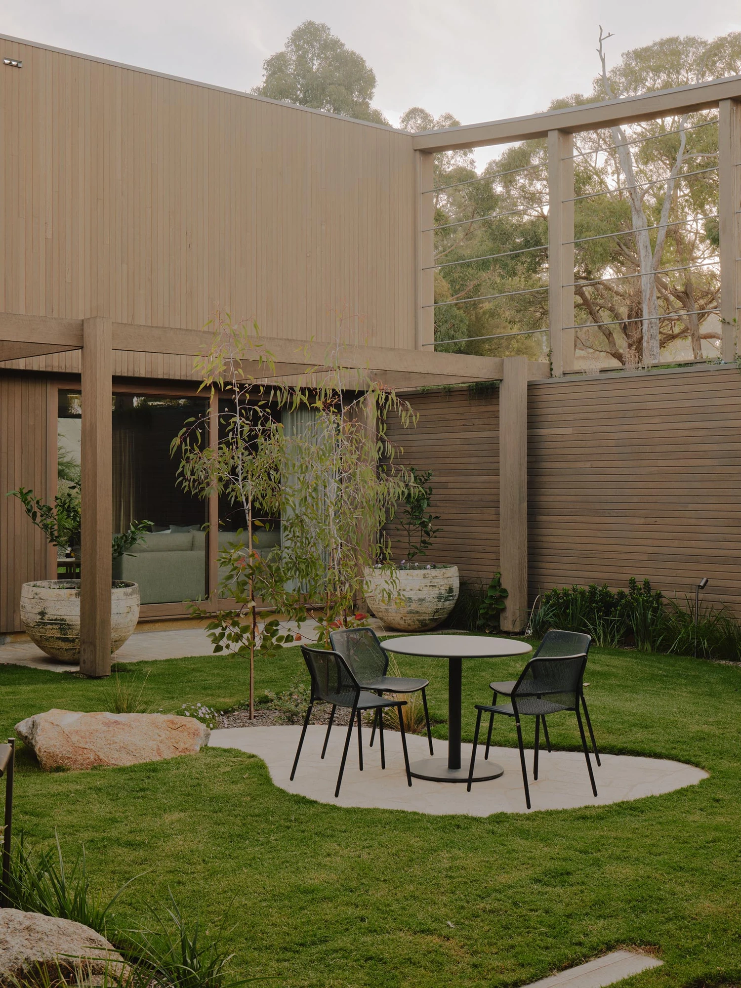The Farm by Wolveridge Architects, photographed by Jack Carlin — A grassed courtyard holds a black cafe table and two chairs on a pale paved circle, weathered urn planters and a young eucalyptus beside a sliding glass wall onto the living room, a cable…