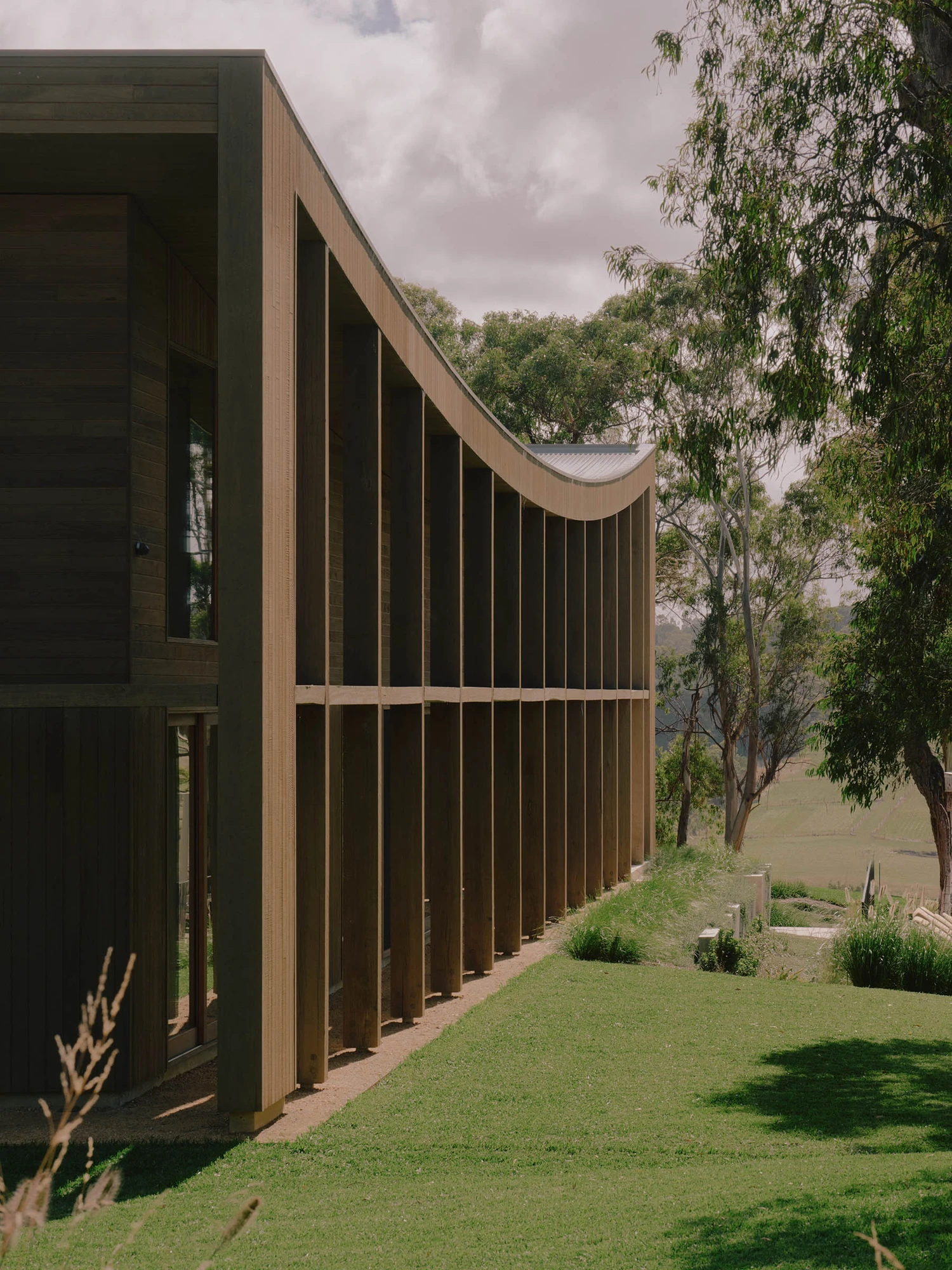The Farm by Wolveridge Architects, photographed by Jack Carlin — The house's curved roofline sweeps down into a tall colonnade of vertical timber posts, dark glazing set behind, above a green lawn that falls away to gums and distant pasture.