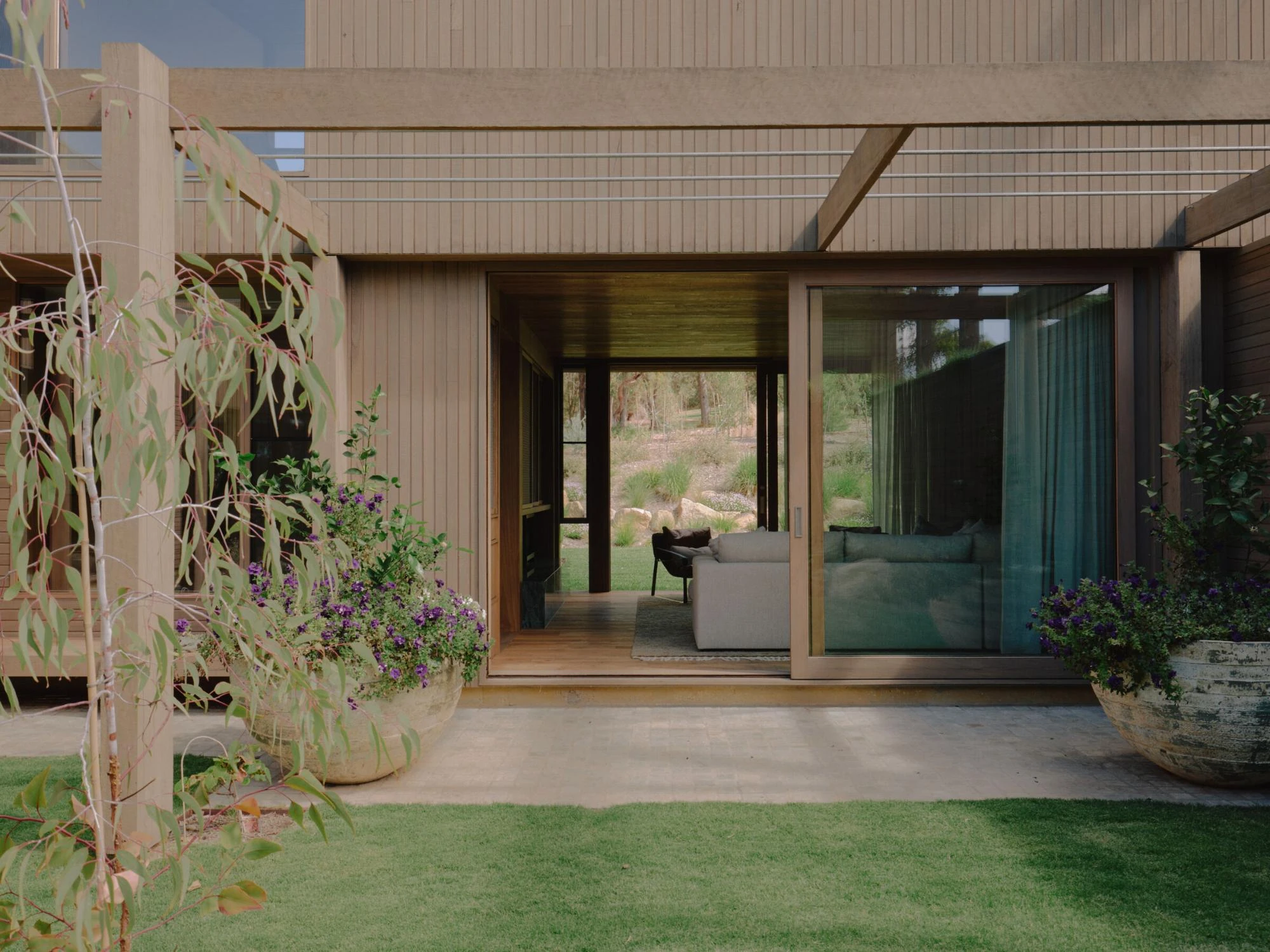 The Farm by Wolveridge Architects, photographed by Jack Carlin — Under a timber pergola a sliding glass wall opens a living room with a grey sofa onto a stone-paved terrace, purple-flowering plants in aged urns to either side and lawn beyond.