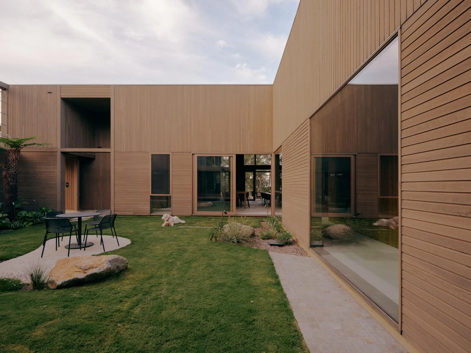 The Farm by Wolveridge Architects, photographed by Jack Carlin — An L-shaped courtyard wrapped in timber cladding, a white dog resting on the lawn, a black table and chairs on a paved circle and sliding glass revealing the dining room through the house.