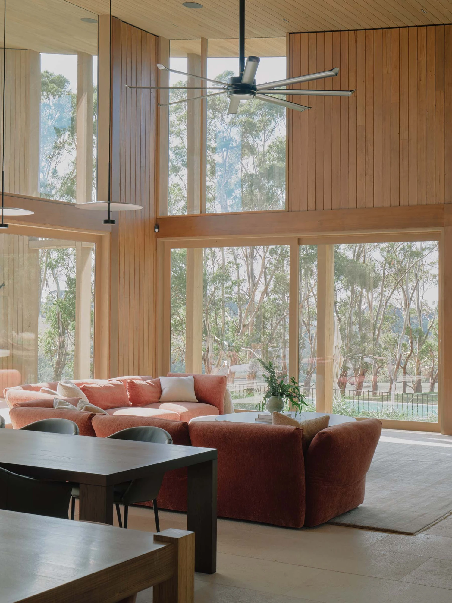 The Farm by Wolveridge Architects, photographed by Jack Carlin — A curved terracotta sectional gathers before a wall of glass looking into the gums, the timber-lined ceiling and ceiling fan overhead, a dark dining table in the foreground.