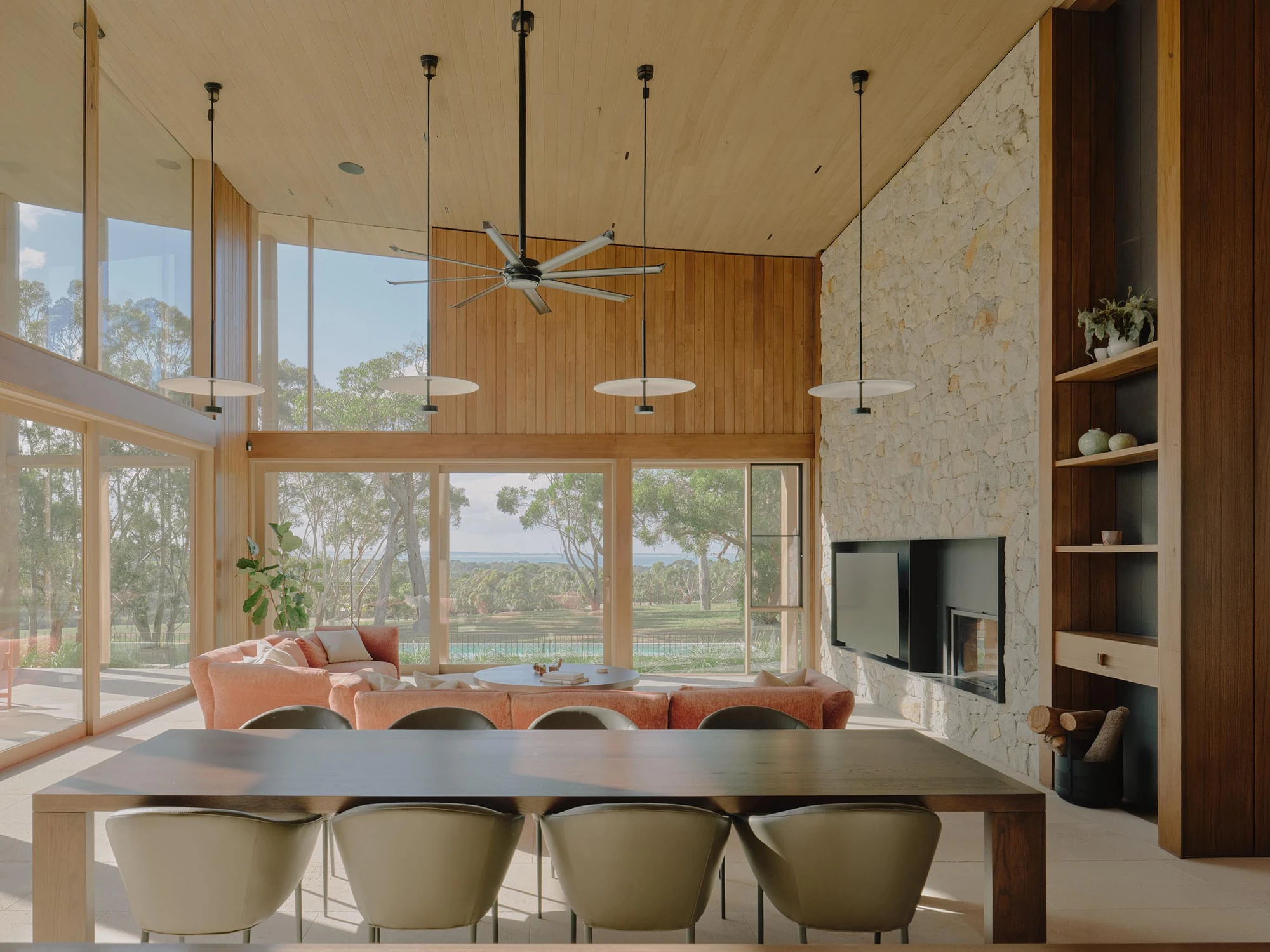 The Farm by Wolveridge Architects, photographed by Jack Carlin — The open living and dining room sits beneath a raked timber ceiling and a wide ceiling fan, a green-veined stone chimney breast holding an inset fire beside timber shelving, a curved…