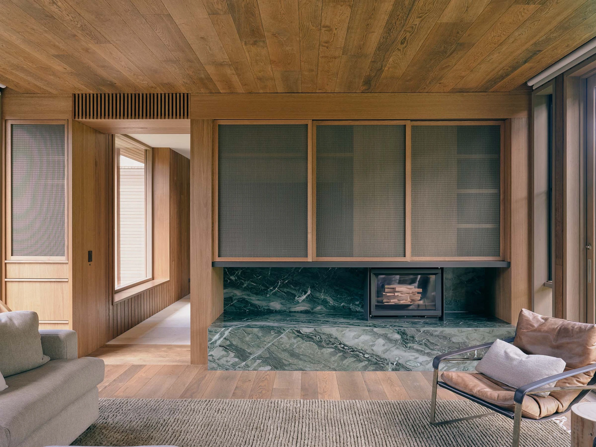 The Farm by Wolveridge Architects, photographed by Jack Carlin — A sitting room lined in timber, a green-veined stone hearth with a wood-burner set below mesh-fronted cabinetry, a grey sofa and a tan leather sling chair on a ribbed rug.