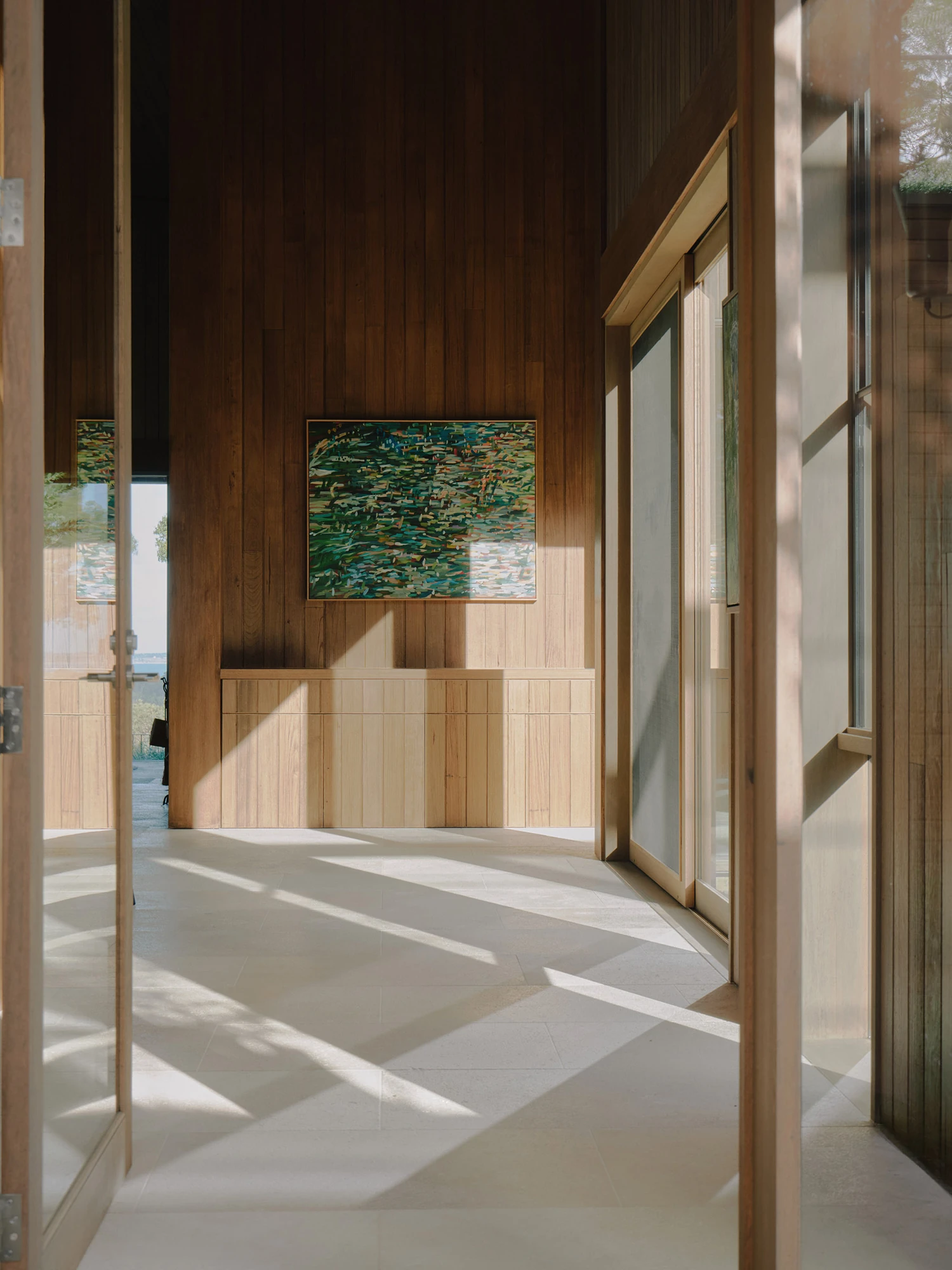 The Farm by Wolveridge Architects, photographed by Jack Carlin — An entry hall floored in pale stone, its timber-clad walls hung with a green landscape painting above a low fitted cabinet, sunlight cast in long bars across the floor.