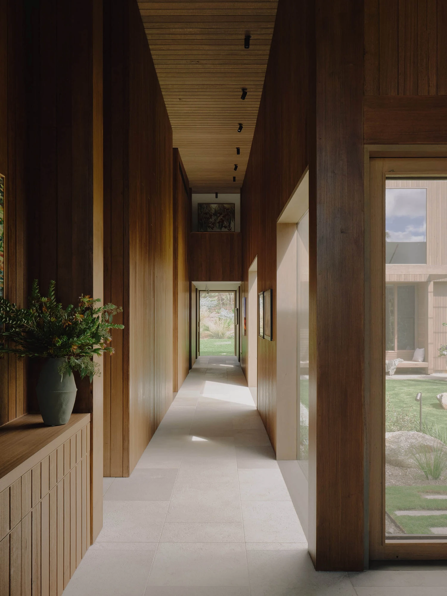 The Farm by Wolveridge Architects, photographed by Jack Carlin — A timber-lined corridor over pale stone, a green vase of native foliage on a low cabinet, framed art on the wall and daylight opening to garden at either end.
