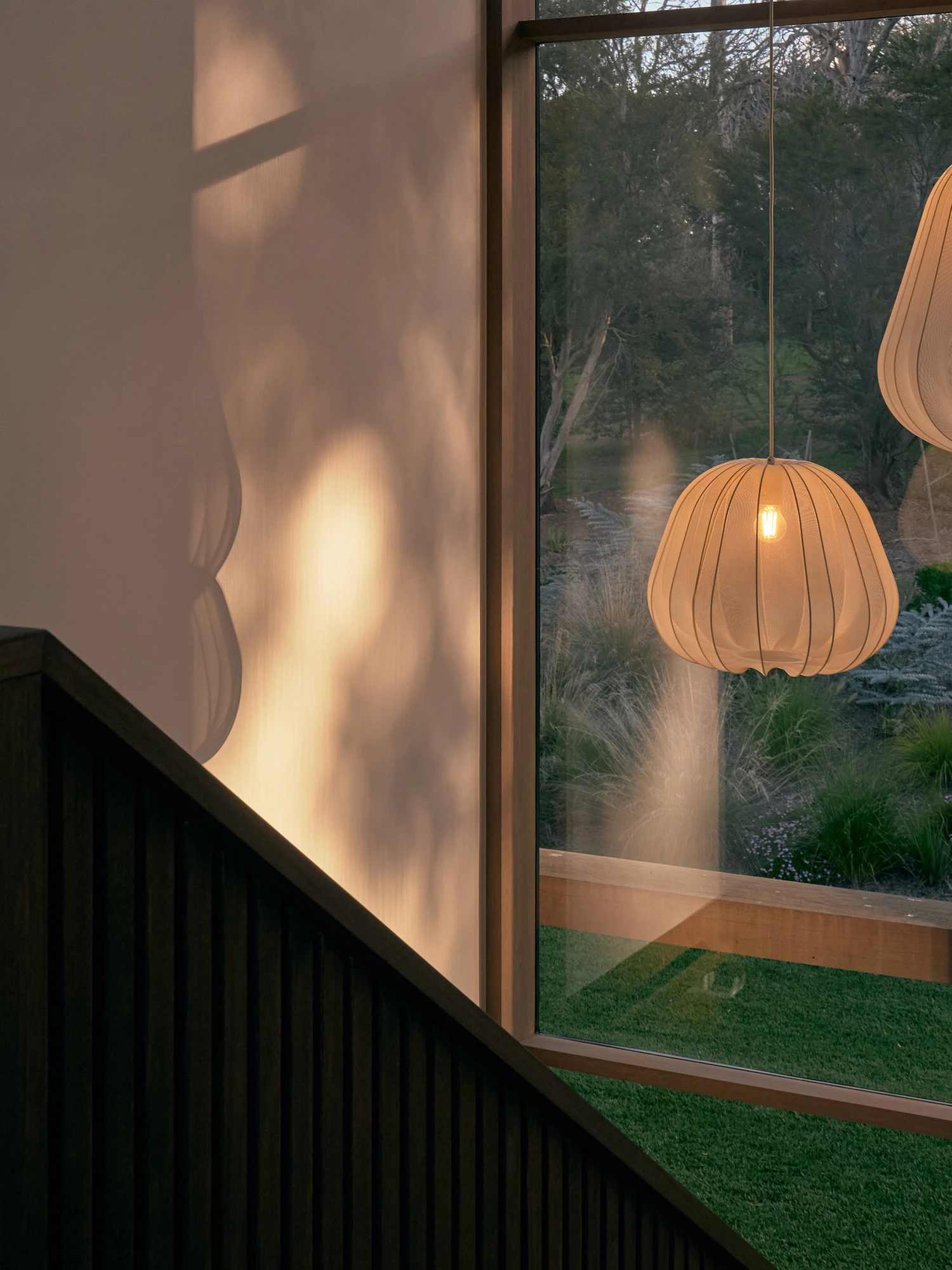 The Farm by Wolveridge Architects, photographed by Jack Carlin — An upper-level corner where dappled light falls on a cream wall above a dark battened balustrade, two gathered-fabric pendant lights hanging at a large window onto the garden.