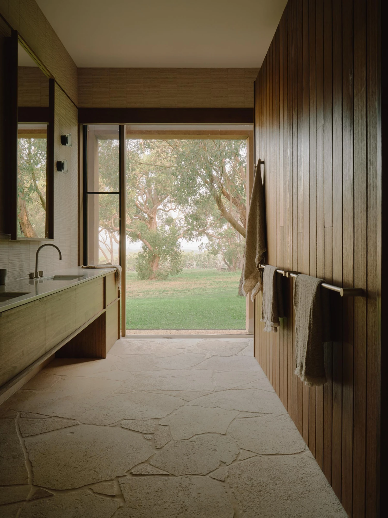 The Farm by Wolveridge Architects, photographed by Jack Carlin — A bathroom floored in crazy-paved stone, a timber-clad wall hung with towels, a stone vanity with twin basins and a black tap, a full-height glazed door open to gums and lawn.