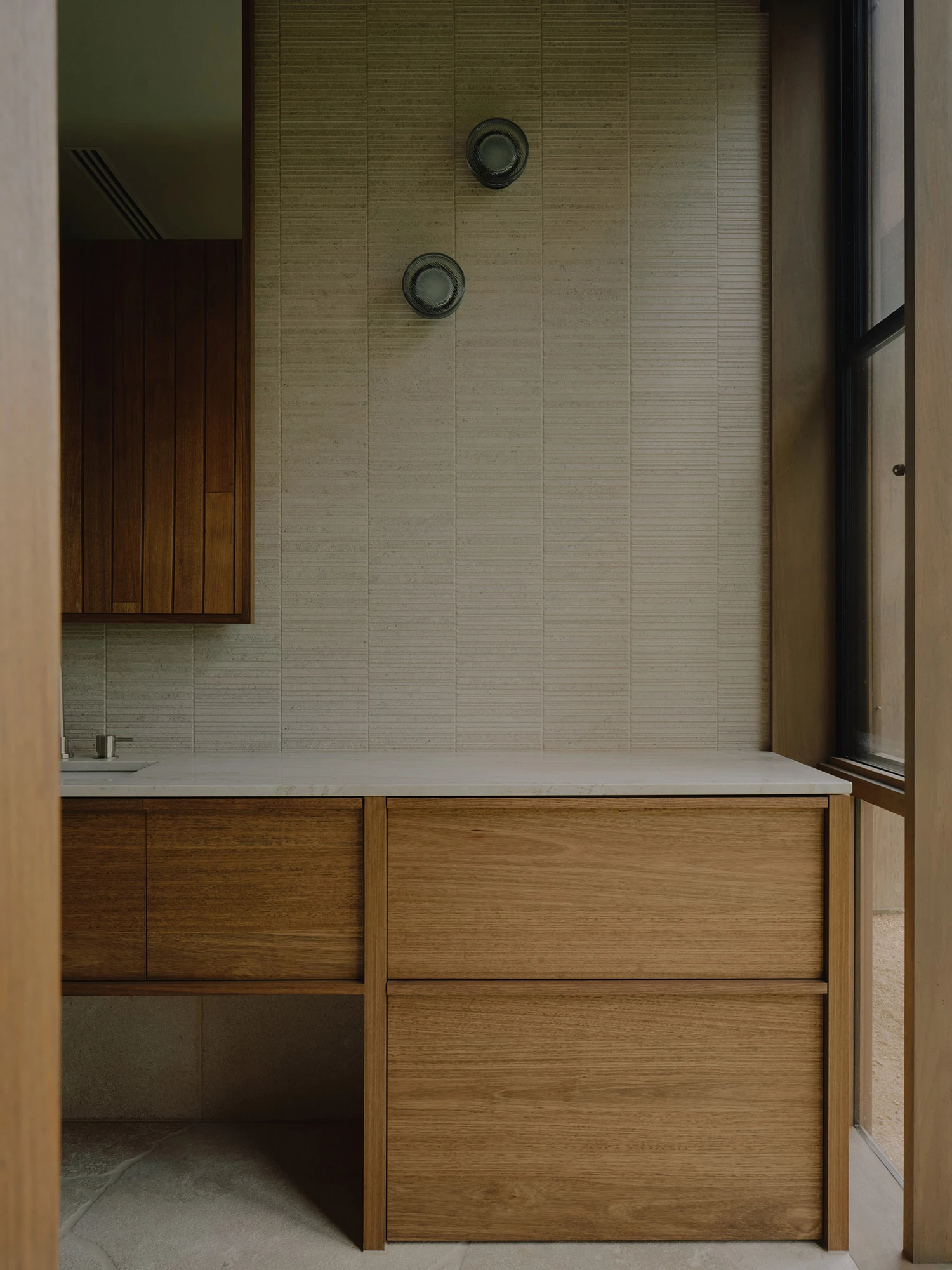 The Farm by Wolveridge Architects, photographed by Jack Carlin — A vanity set against a ribbed pale-tiled wall with two round glass wall lights, timber cabinetry below a stone top and a window to one side.
