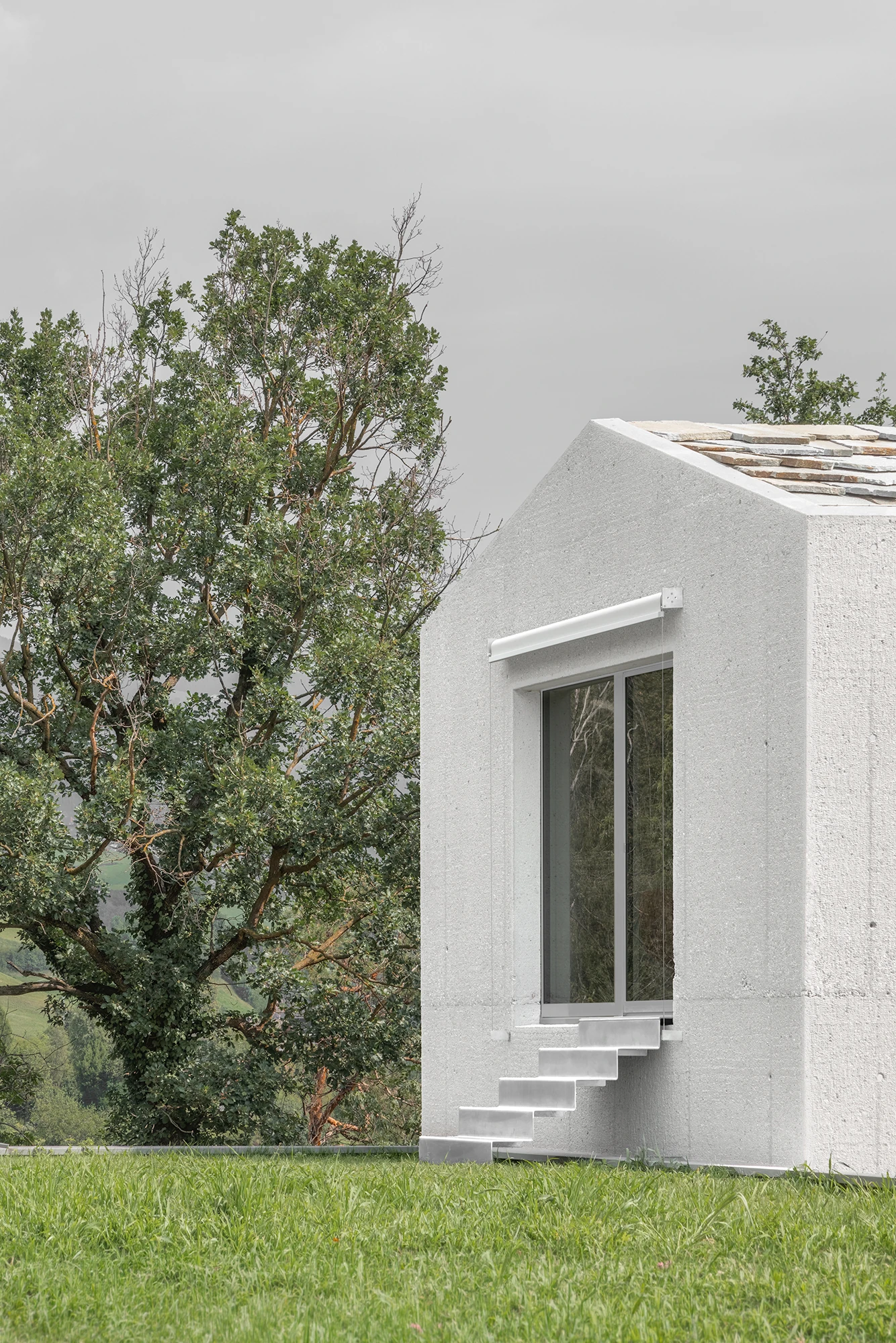 Val Mira by Andreas Gruber Architects, photographed by Gustav Willeit — A folded metal stair climbs the concrete flank of a gabled tower to a sliding window, stone slabs edging the roof, a large tree and green valley alongside.