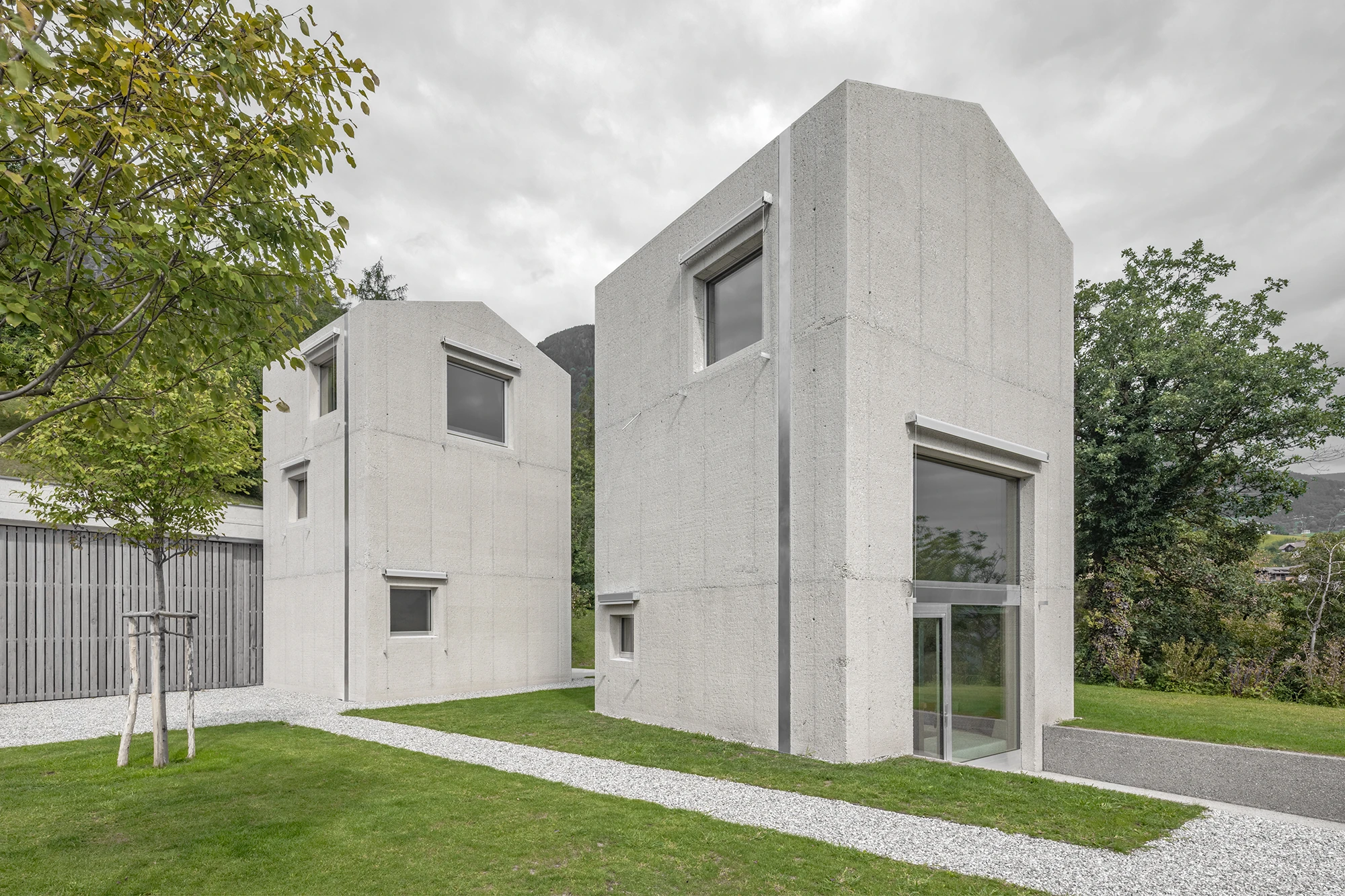 Val Mira by Andreas Gruber Architects, photographed by Gustav Willeit — Two concrete towers with faceted gabled caps meet across a gravel path, a young staked tree and grey timber fence to the left and mountains fading behind.