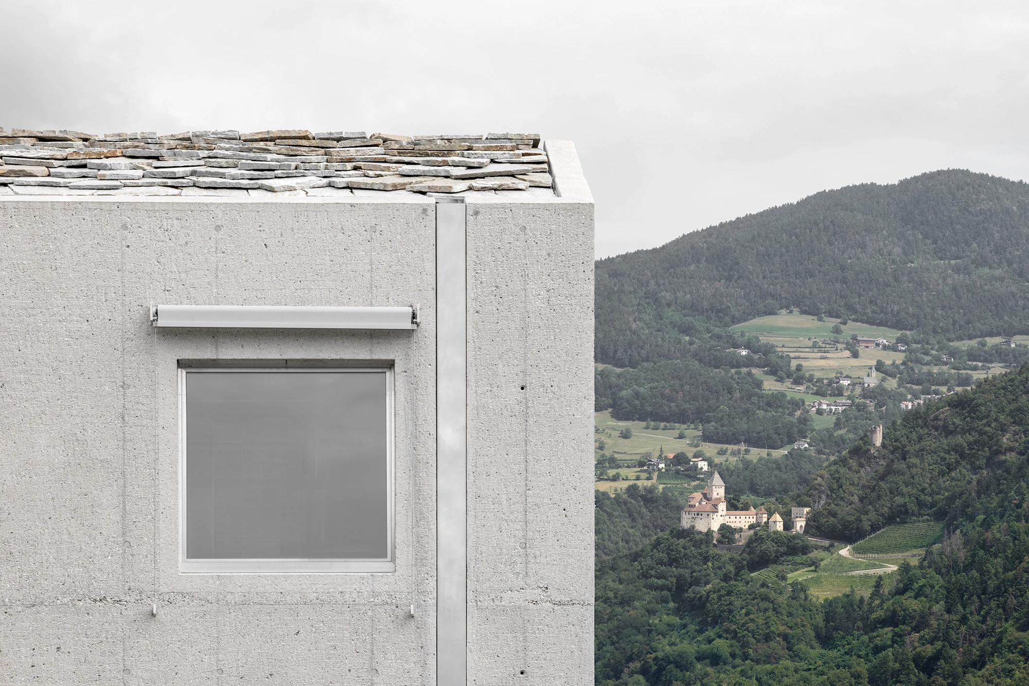 Val Mira by Andreas Gruber Architects, photographed by Gustav Willeit — Corner detail of a concrete tower, stone slabs stacked along the pitched roof edge above a shaded square window, a hilltop castle across the wooded valley.