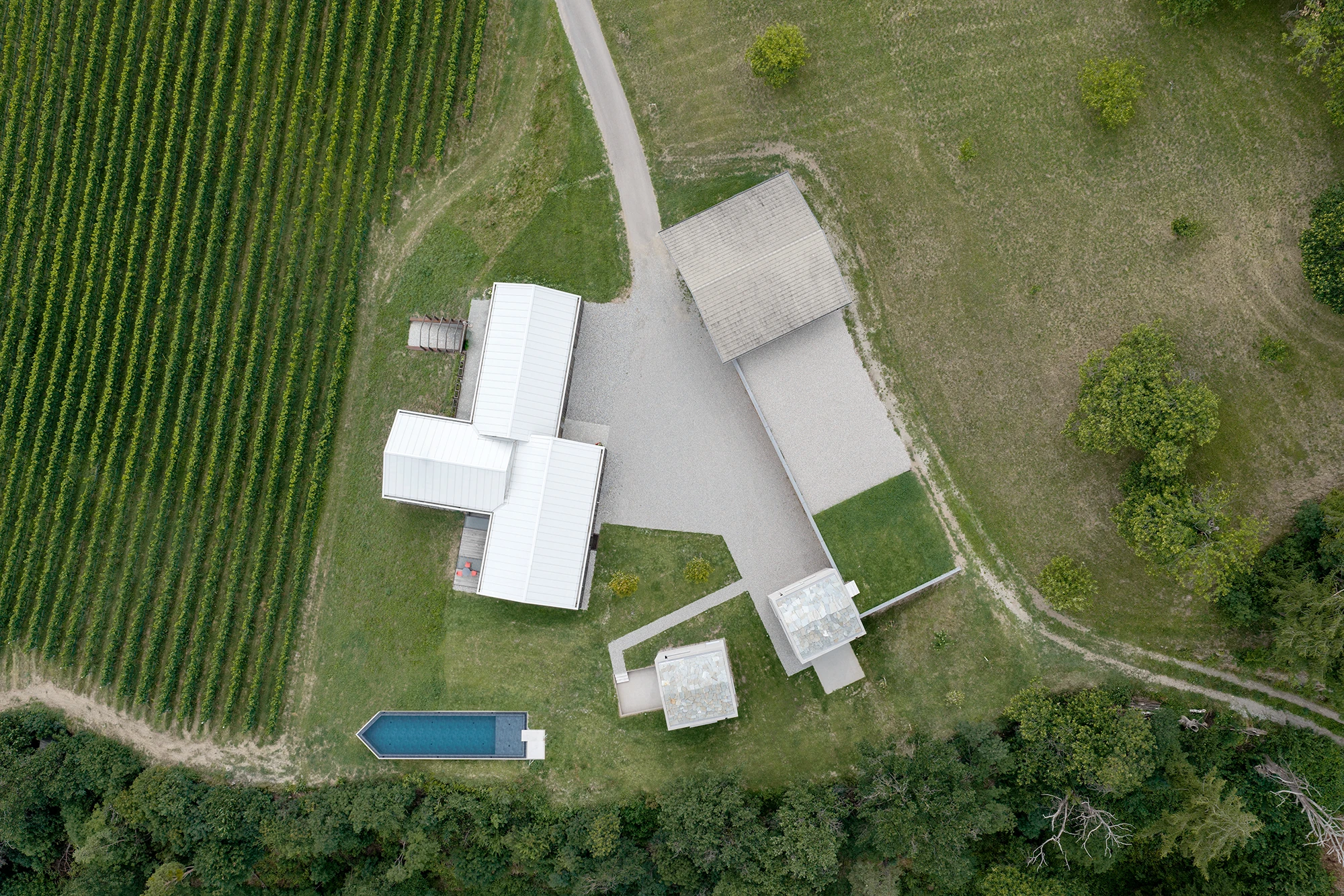 Val Mira by Andreas Gruber Architects, photographed by Gustav Willeit — Aerial of the full ensemble: the white-roofed timber house and outbuildings around a gravel court with the two stone-roofed towers and the slim pool below, vineyard rows to one side.