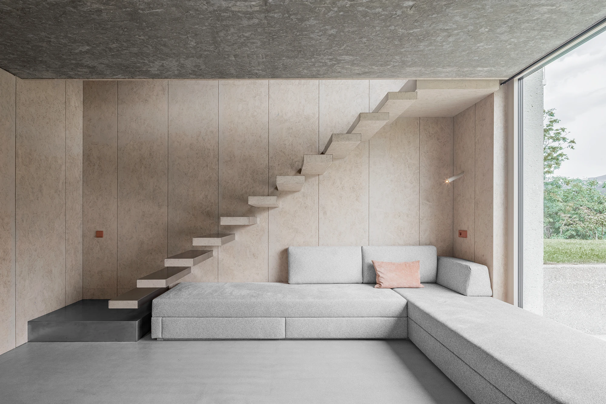 Val Mira by Andreas Gruber Architects, photographed by Gustav Willeit — Wood-lined living room with a pale cantilevered staircase floating up the back wall above a low grey sectional, concrete ceiling and full-height glazing to the right.