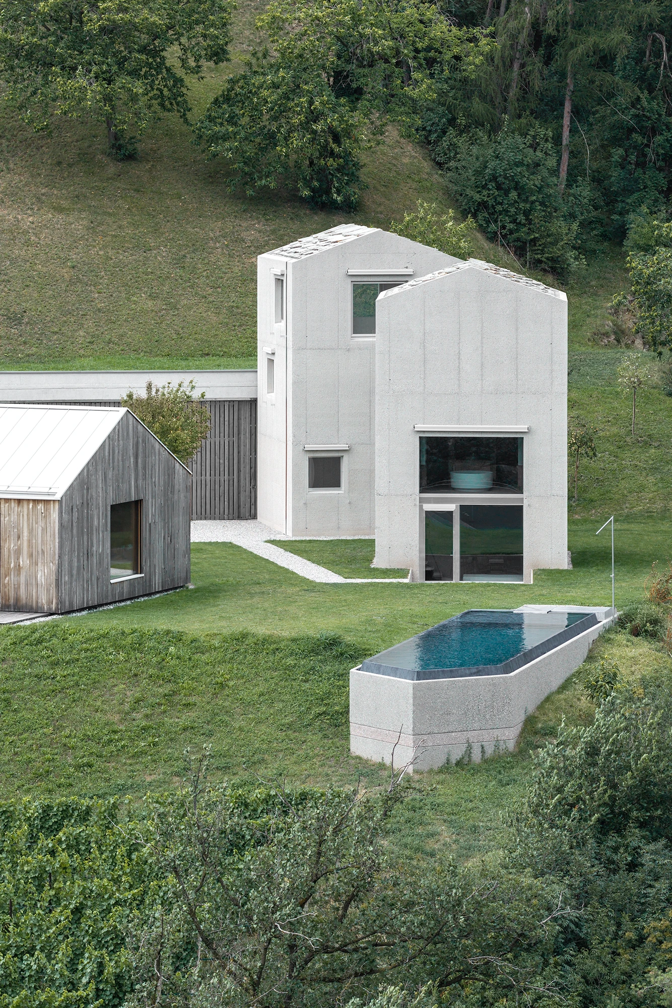 Val Mira by Andreas Gruber Architects, photographed by Gustav Willeit — Vertical view of the two stone-roofed concrete towers on the hillside, a timber outbuilding at left and a long dark-rimmed pool set into the meadow below.