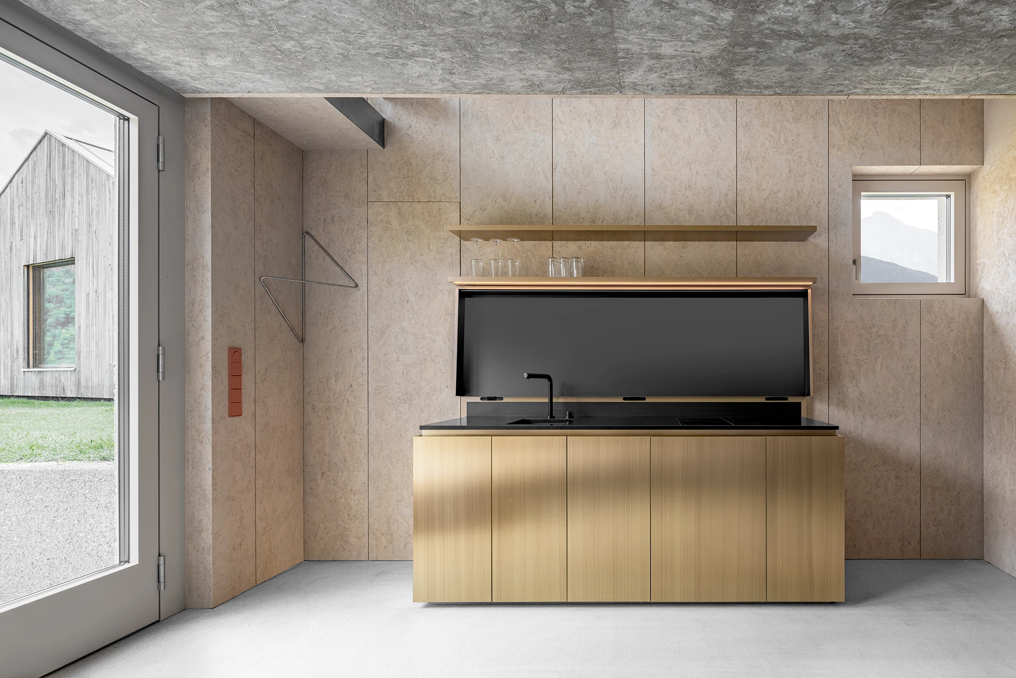 Val Mira by Andreas Gruber Architects, photographed by Gustav Willeit — Brass kitchen block with a black stone counter and folding black backsplash against wood-panelled walls, a glazed door framing the timber building outside.