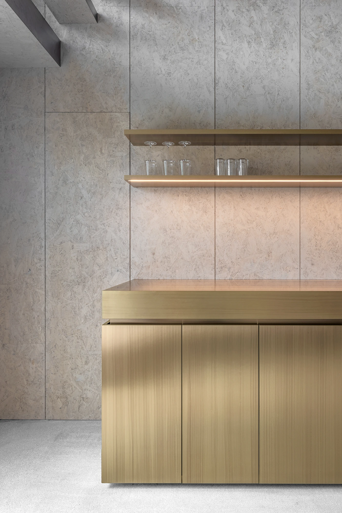 Val Mira by Andreas Gruber Architects, photographed by Gustav Willeit — Close view of the brushed-brass kitchen cabinet and a lit wood shelf lined with glasses against the wood-panelled wall.