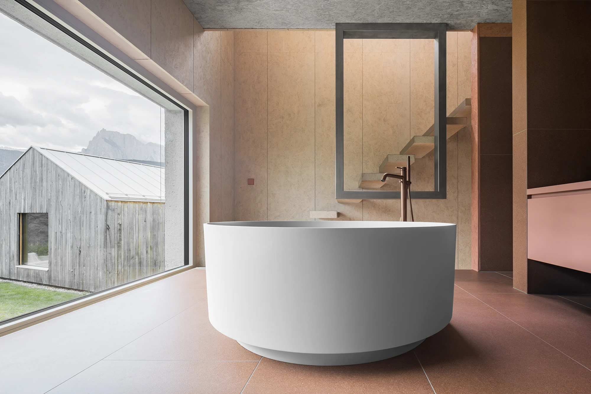 Val Mira by Andreas Gruber Architects, photographed by Gustav Willeit — Round freestanding bathtub on rust-red floor tiles before a wide window, wood-panelled walls and a framed mirror reflecting the floating stair, a copper tap alongside.