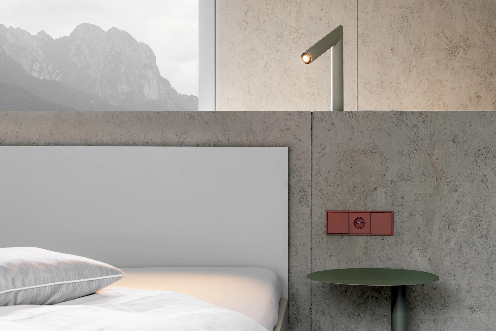 Val Mira by Andreas Gruber Architects, photographed by Gustav Willeit — Bedroom corner with white bed linen, a green reading lamp and side table against wood-panelled walls, a terracotta socket plate and a misty mountain view through the window.