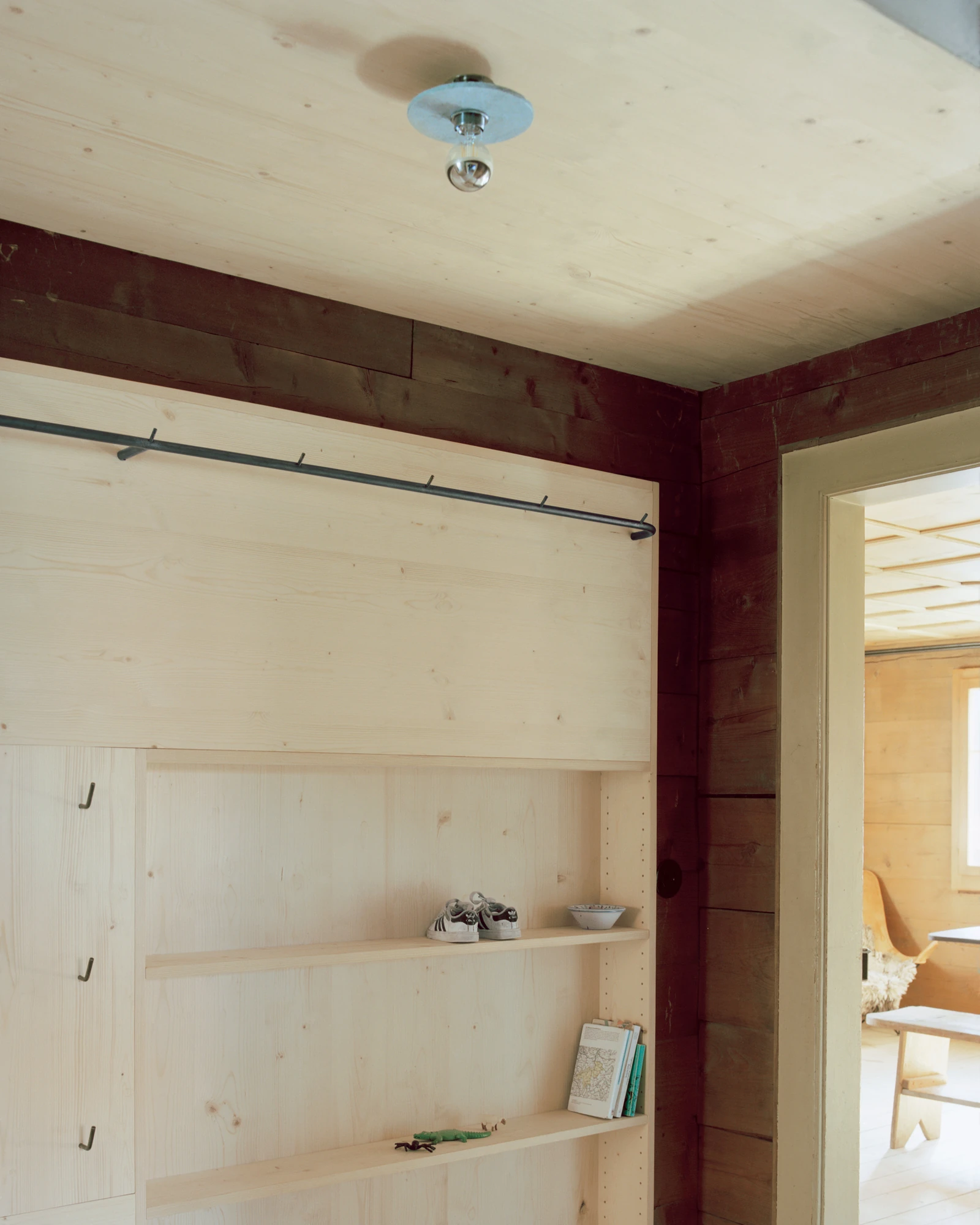 Via Seneria by Dario Wohler, photographed by Willem Pab — Built-in pale timber shelving and a bracketed rail set into a wall of dark red-brown boards, holding small shoes and a folded map, a lit room visible through the doorway to the right.