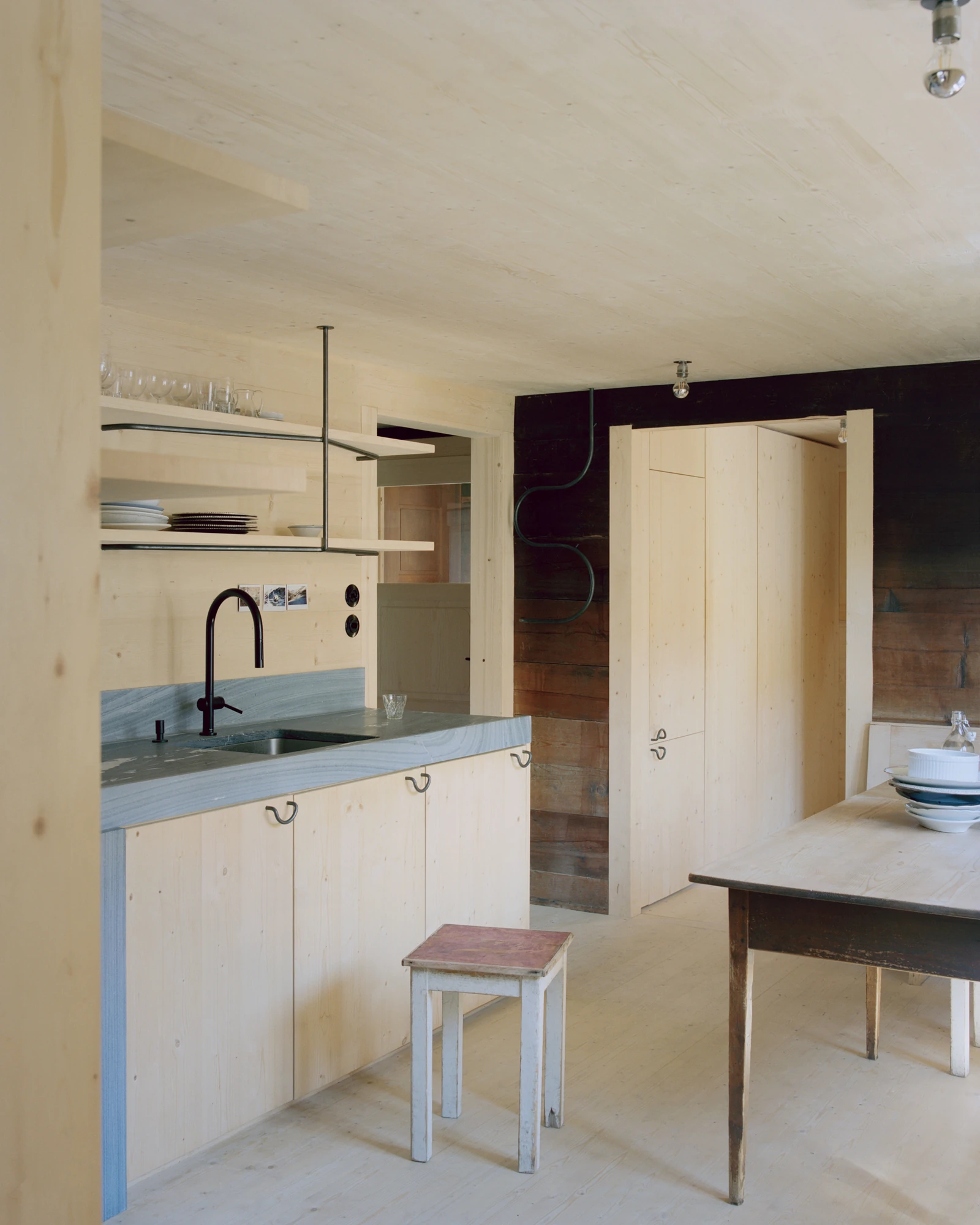 Via Seneria by Dario Wohler, photographed by Willem Pab — A compact kitchen with pale timber cabinets, a grey veined stone counter and a black gooseneck tap, set against a dark timber wall beside a light cupboard.
