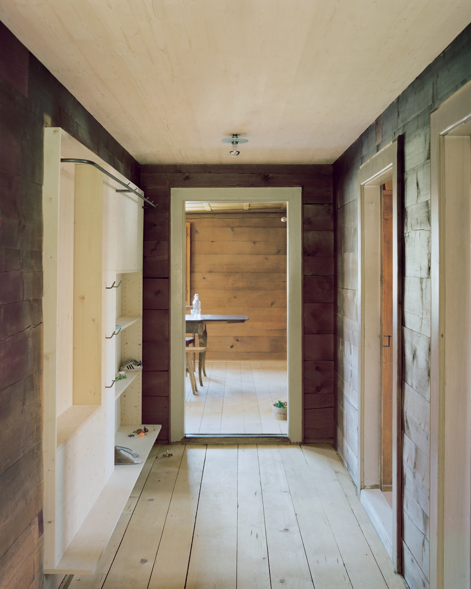 Via Seneria by Dario Wohler, photographed by Willem Pab — A corridor of pale plank flooring runs between dark timber walls toward a bright doorway framing a table and chair, with a built-in bench and shelves along the left side.