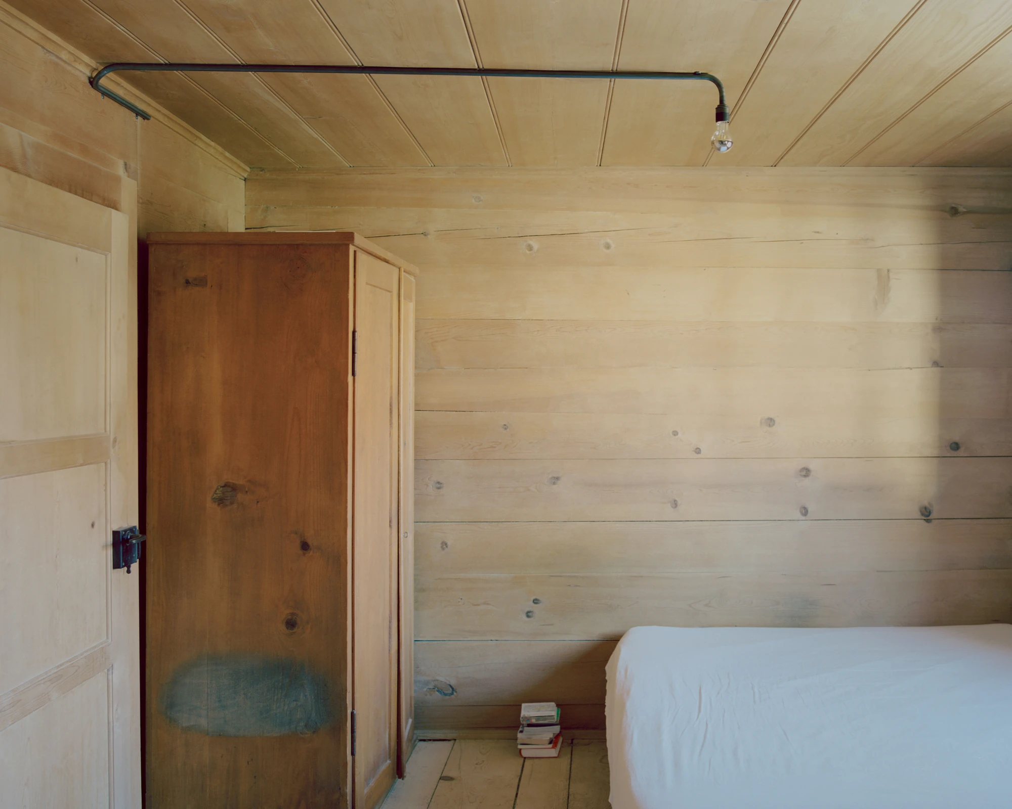 Via Seneria by Dario Wohler, photographed by Willem Pab — A bedroom lined in pale honey-toned timber holds a wooden wardrobe and a made bed, with books stacked on the plank floor and a bent metal conduit carrying a bare bulb across the ceiling.