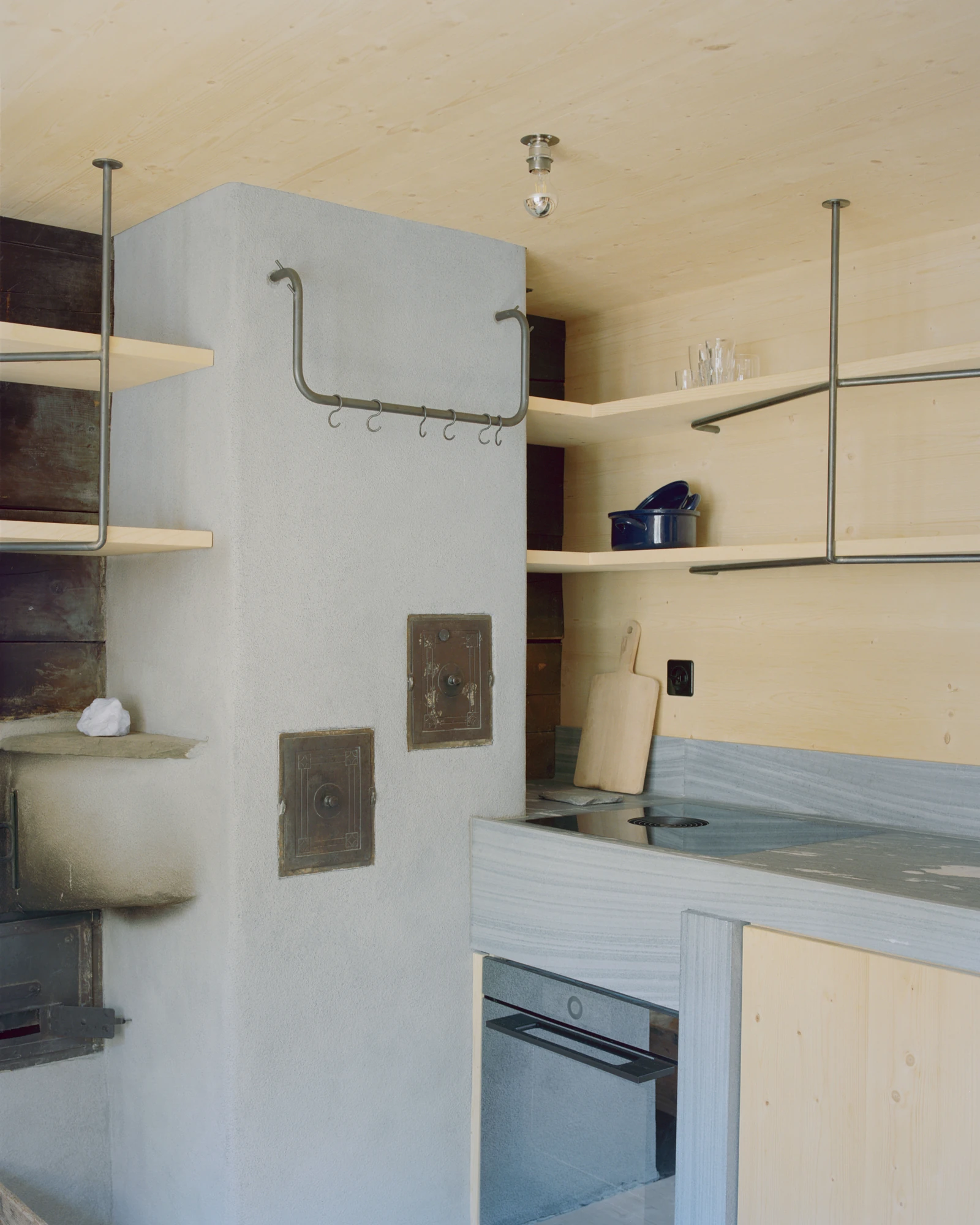 Via Seneria by Dario Wohler, photographed by Willem Pab — A rendered grey chimney mass fitted with two cast-iron oven doors stands beside a stone counter and hob, ringed by metal hanging rails and pale timber shelves.