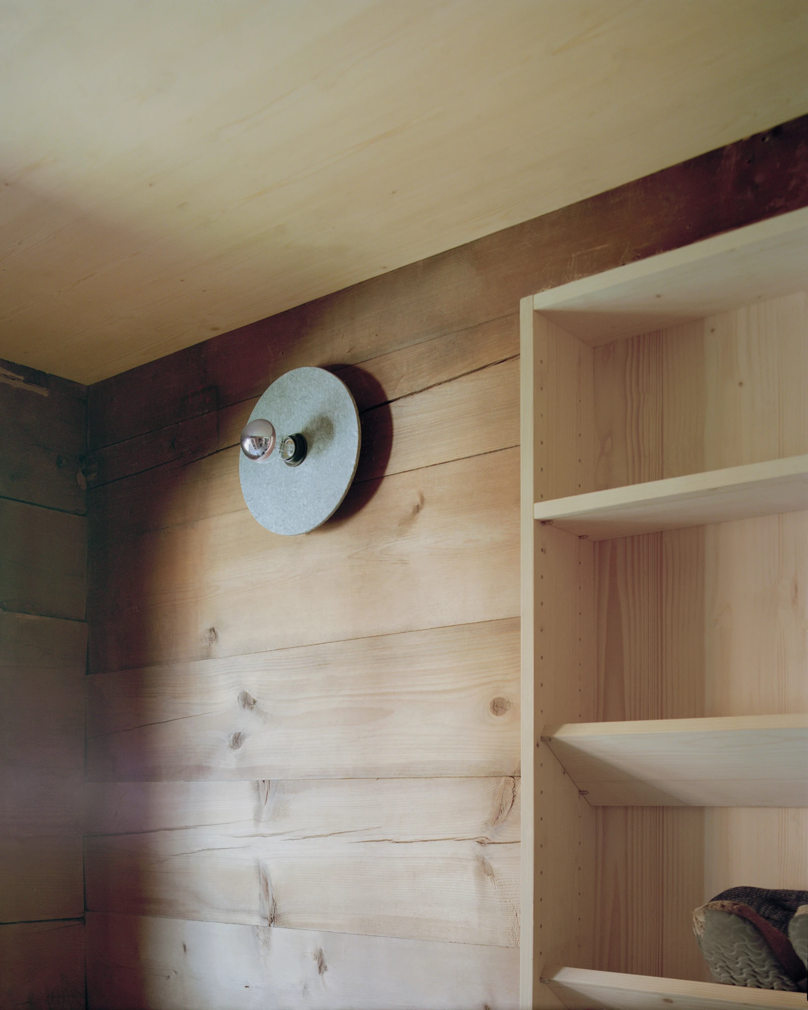 Via Seneria by Dario Wohler, photographed by Willem Pab — A round grey stone disc light holding a mirrored bulb is mounted on a wall of pale timber boards, beside the open edge of a shelving unit.