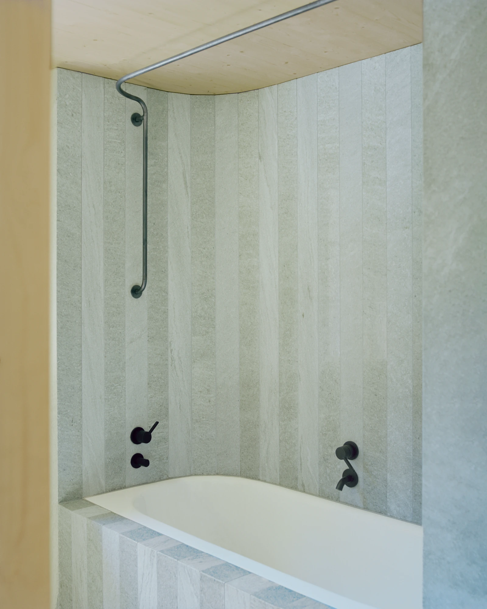 Via Seneria by Dario Wohler, photographed by Willem Pab — A bathtub set into a niche lined in pale grey stone, with matte black wall taps and a curved metal shower rail beneath a pale timber ceiling.