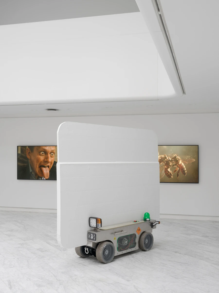 Walls at the World’s End by Louis Morlæ, photographed by David Stjernholm — A single white panel riding a grey four-wheeled cart fills the foreground, two wide video screens on the far wall behind it, one a man with his tongue out and the other a golden…