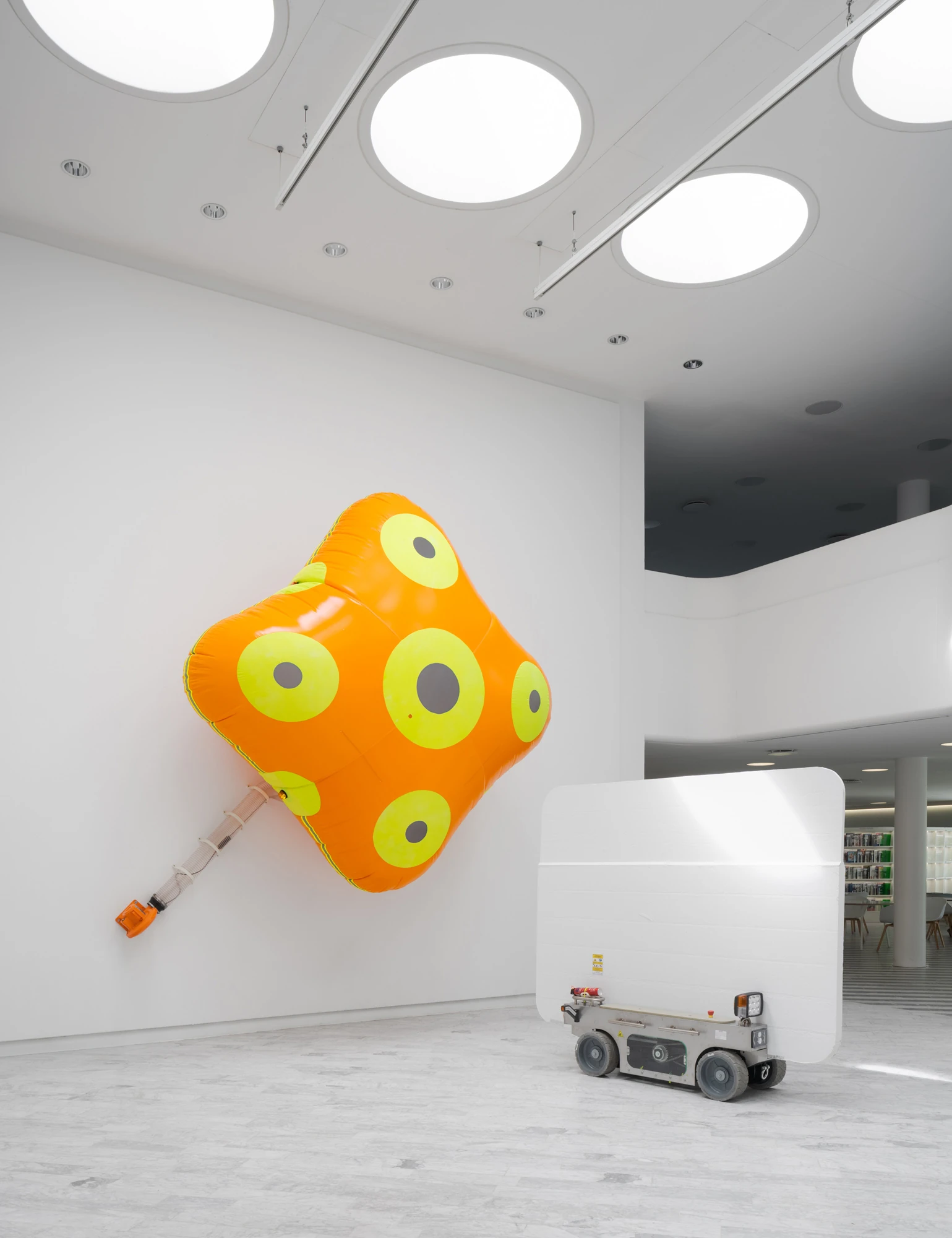 Walls at the World’s End by Louis Morlæ, photographed by David Stjernholm — The many-eyed orange inflatable hangs on a white wall, tubing snaking to its orange blower, while a white panel-cart and a grey robot chassis wait at lower right with the…