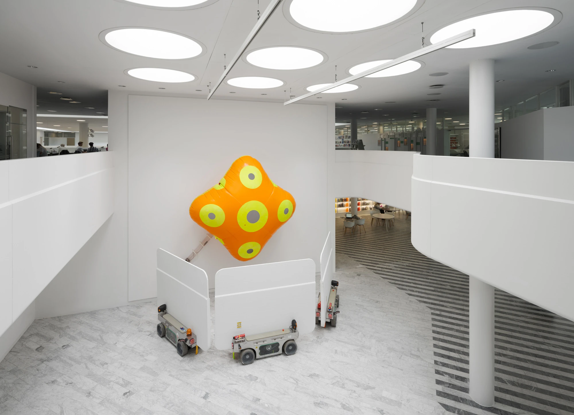 Walls at the World’s End by Louis Morlæ, photographed by David Stjernholm — In the atrium a four-pointed orange inflatable with yellow discs is mounted high on the wall above white panels on carts, a grey column at right and an open void dropping to the…
