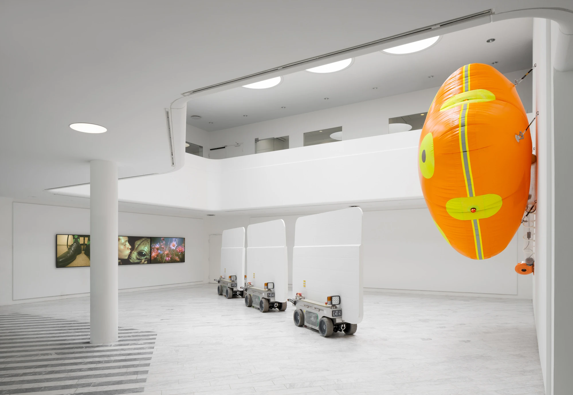 Walls at the World’s End by Louis Morlæ, photographed by David Stjernholm — Seen from a corner, the orange oval inflatable with a yellow-and-grey hazard band clings to the wall in the right foreground while three white panels on carts line up diagonally…
