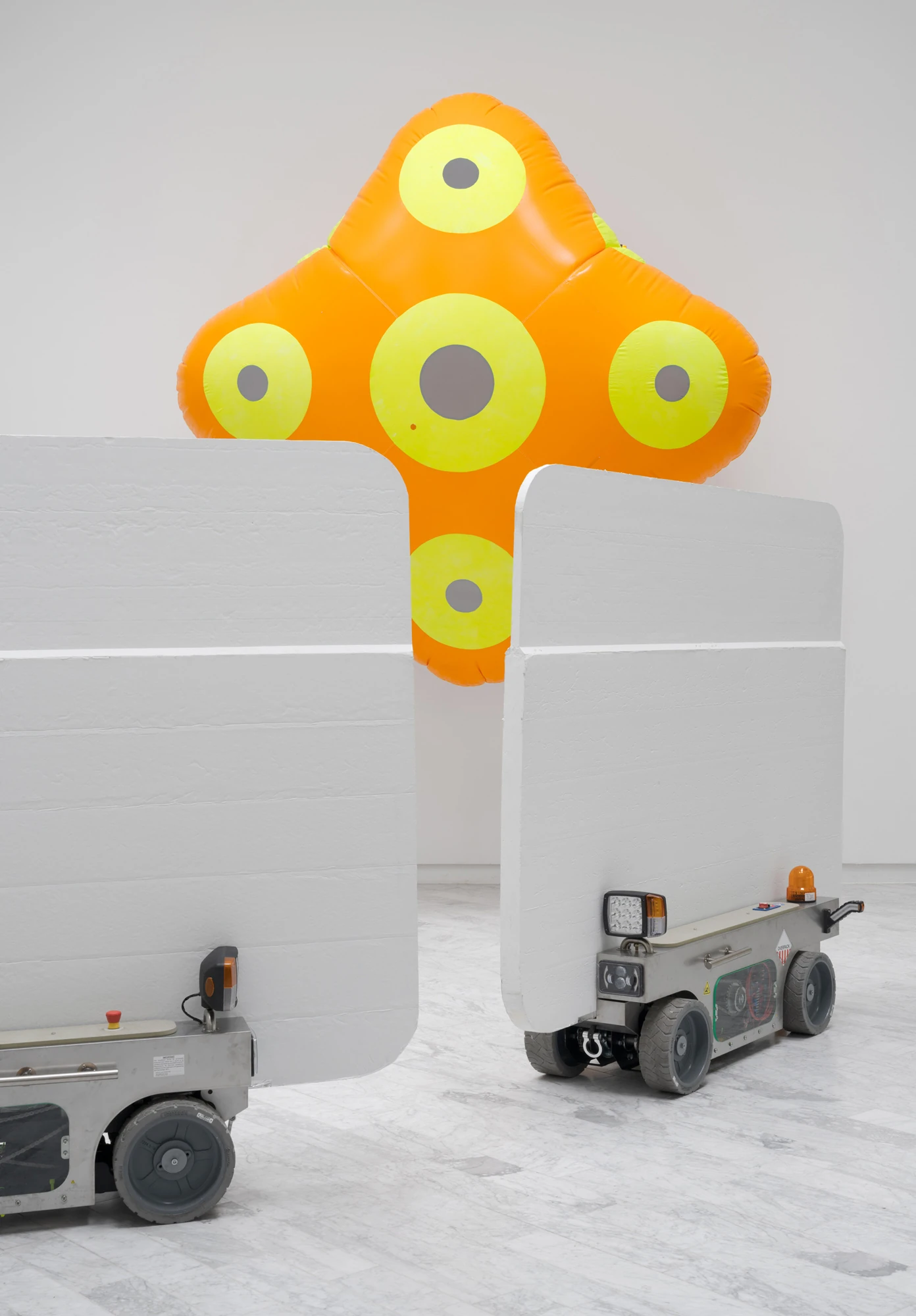 Walls at the World’s End by Louis Morlæ, photographed by David Stjernholm — Two white panel-carts stand in the foreground as a cross-shaped orange inflatable with yellow discs rises behind them, ceiling rings overhead and marble underfoot.