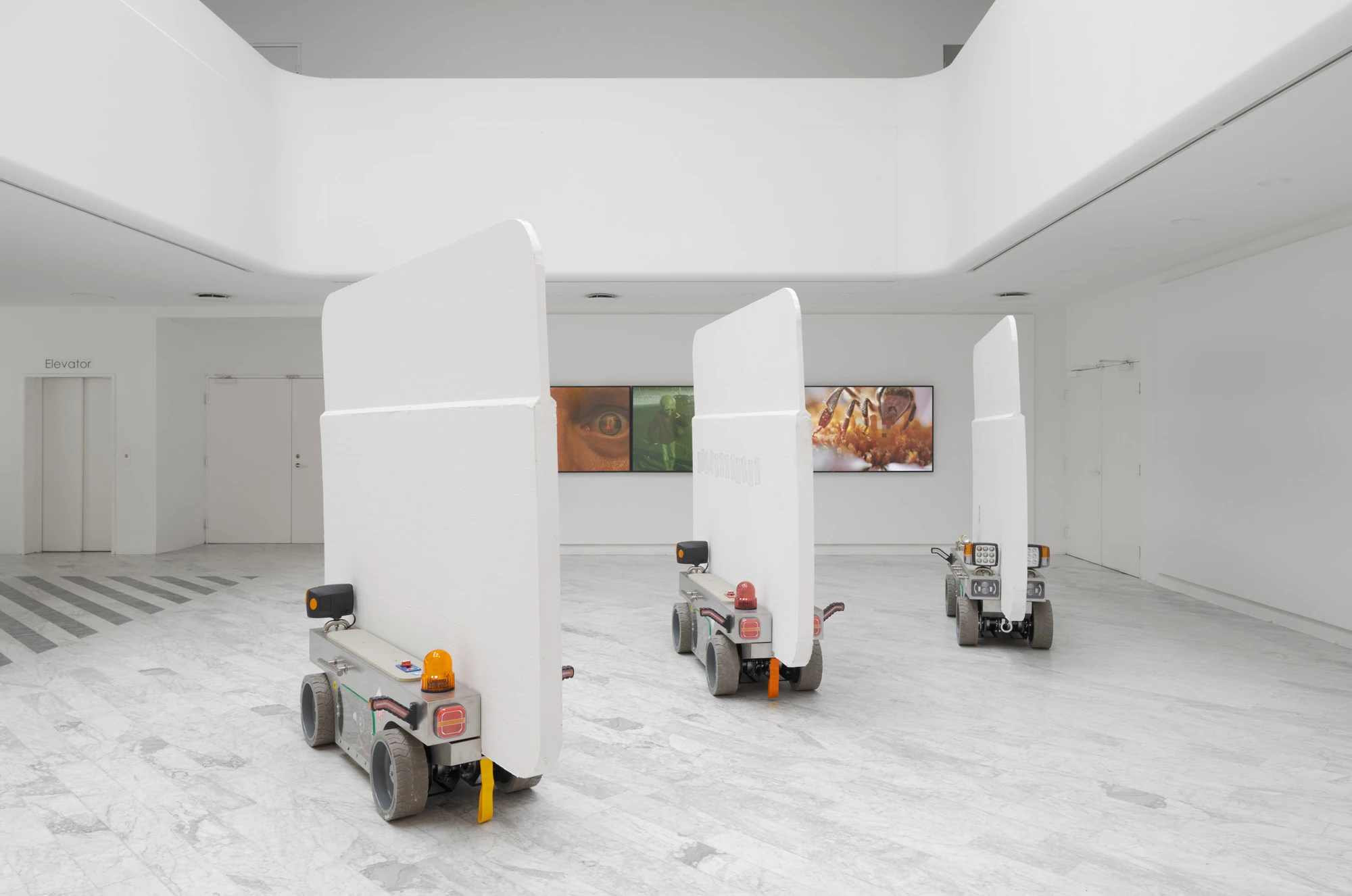 Walls at the World’s End by Louis Morlæ, photographed by David Stjernholm — Three tall white panels ride their carts across the hall, a three-part video on the back wall showing an eye, a green figure and a bee, an Elevator sign on the left door and…