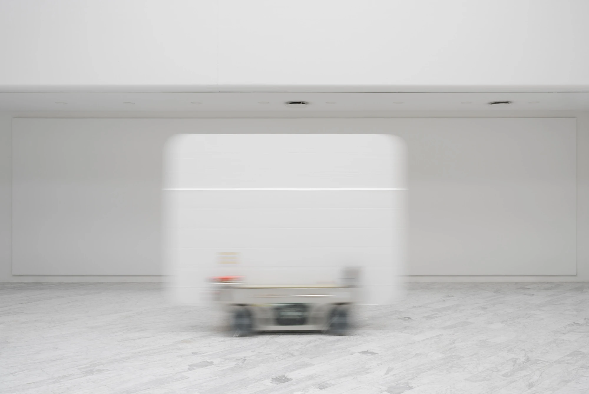 Walls at the World’s End by Louis Morlæ, photographed by David Stjernholm — A white panel on its cart blurs across the frame in motion, dragging its own smear of light past the empty white walls of the hall over marble tiles.
