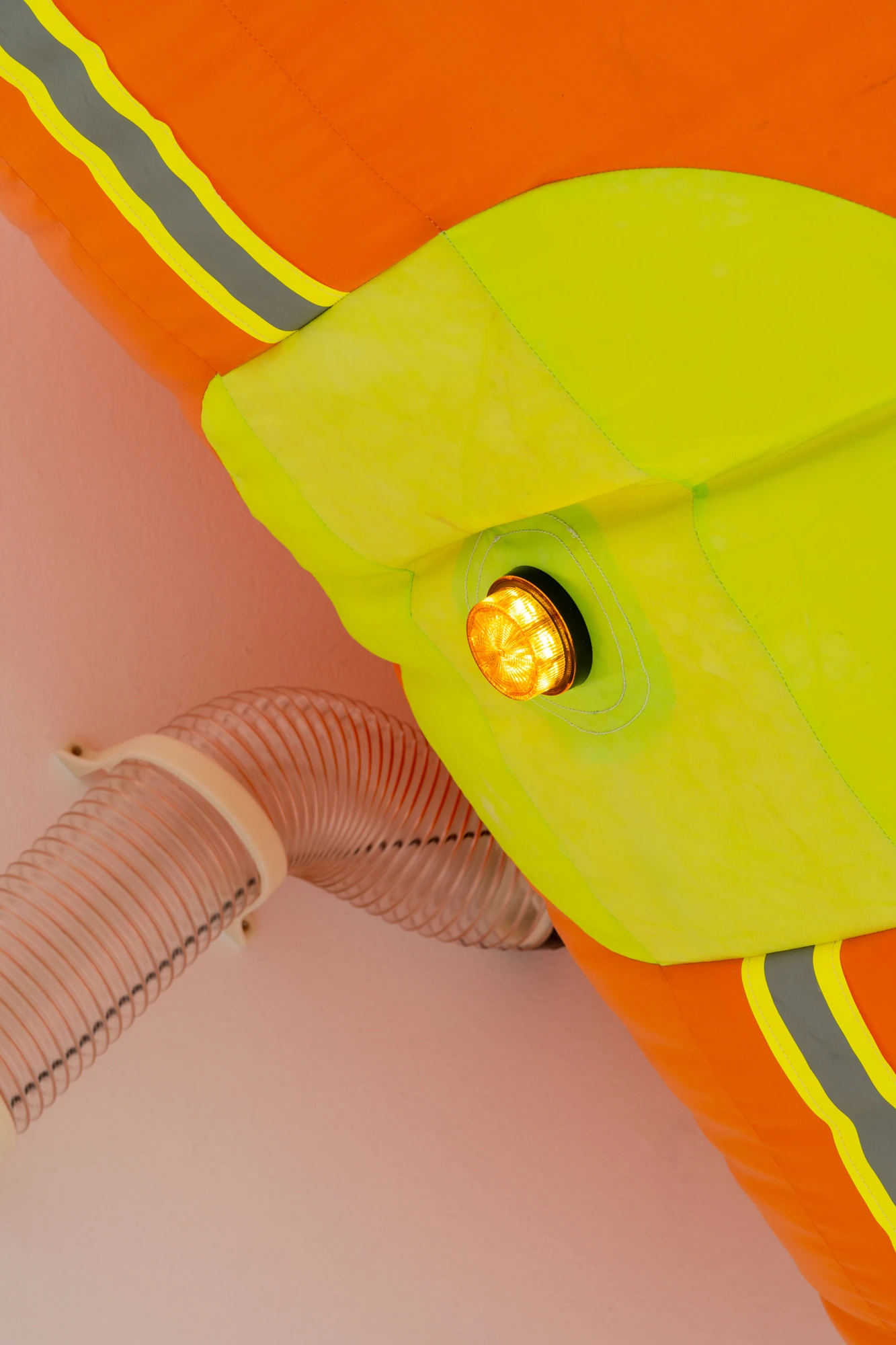 Walls at the World’s End by Louis Morlæ, photographed by David Stjernholm — Extreme close-up of the inflatable, orange fabric seamed with a hazard-yellow band and a yellow panel holding a glowing amber lamp, a ribbed transparent tube curling against a…