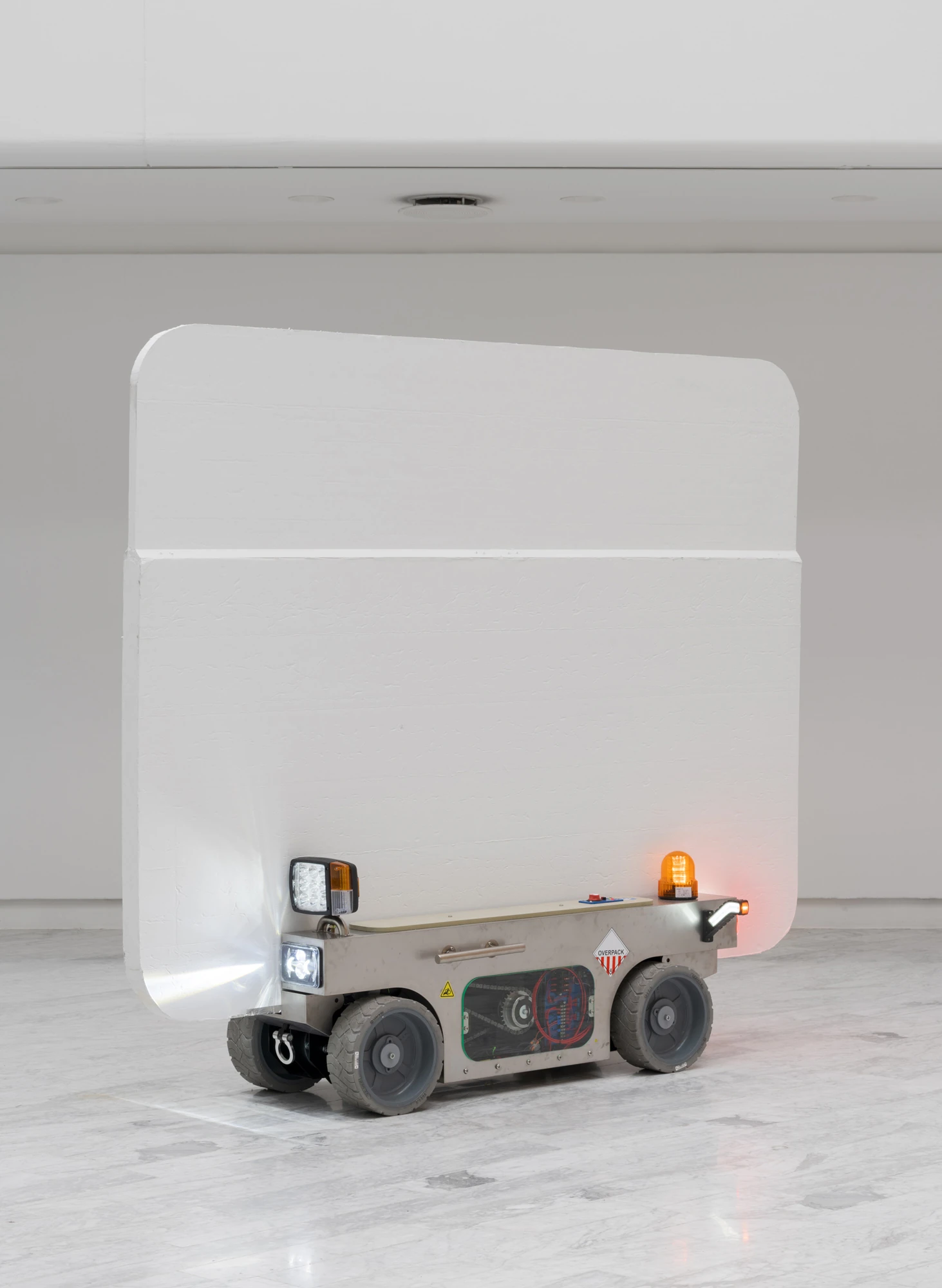 Walls at the World’s End by Louis Morlæ, photographed by David Stjernholm — A single white curved-corner panel on a grey four-wheeled cart faces the camera, headlight and amber beacon lit, alone on the marble floor of the empty white hall.