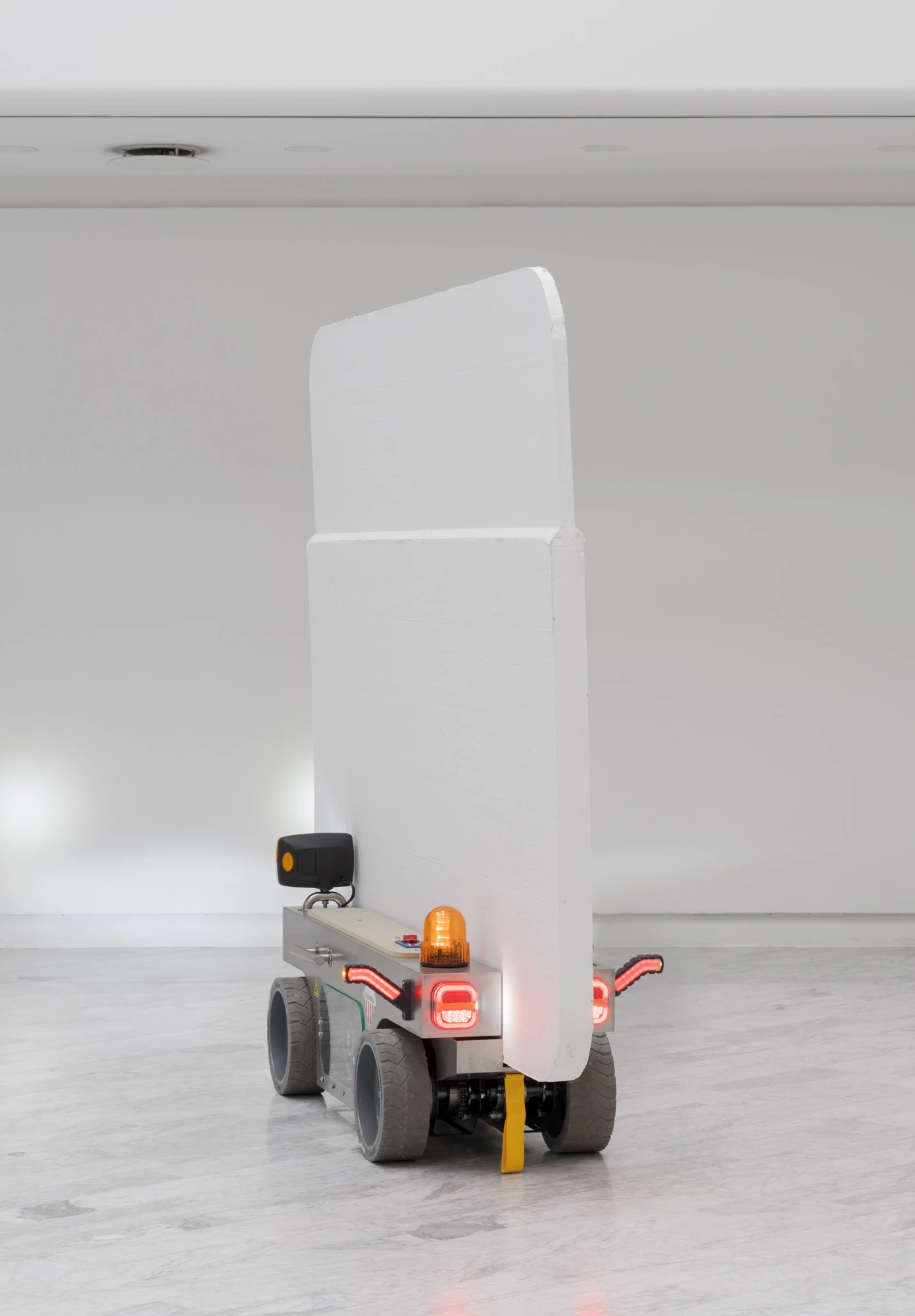 Walls at the World’s End by Louis Morlæ, photographed by David Stjernholm — The white panel-cart seen from behind at an angle, its tall panel rising over a chassis with red tail-lights and an amber beacon lit and a yellow strap hanging loose.