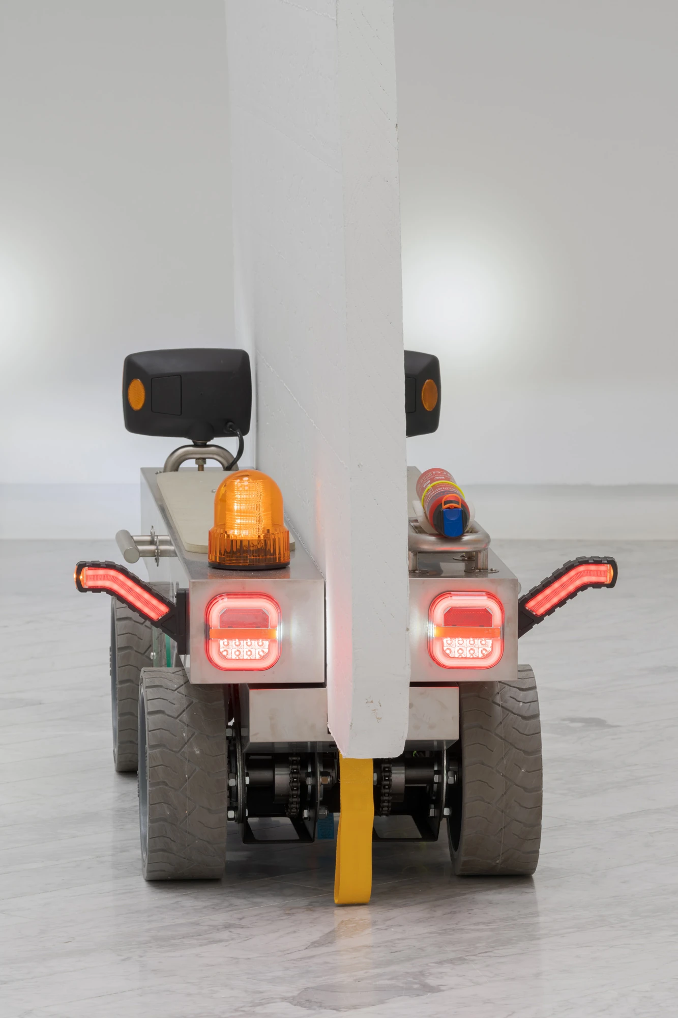 Walls at the World’s End by Louis Morlæ, photographed by David Stjernholm — Rear close-up of a cart nosed against a white column, twin amber beacons and red tail-lights lit, the drive chain and a yellow strap visible below the panel that rises behind.