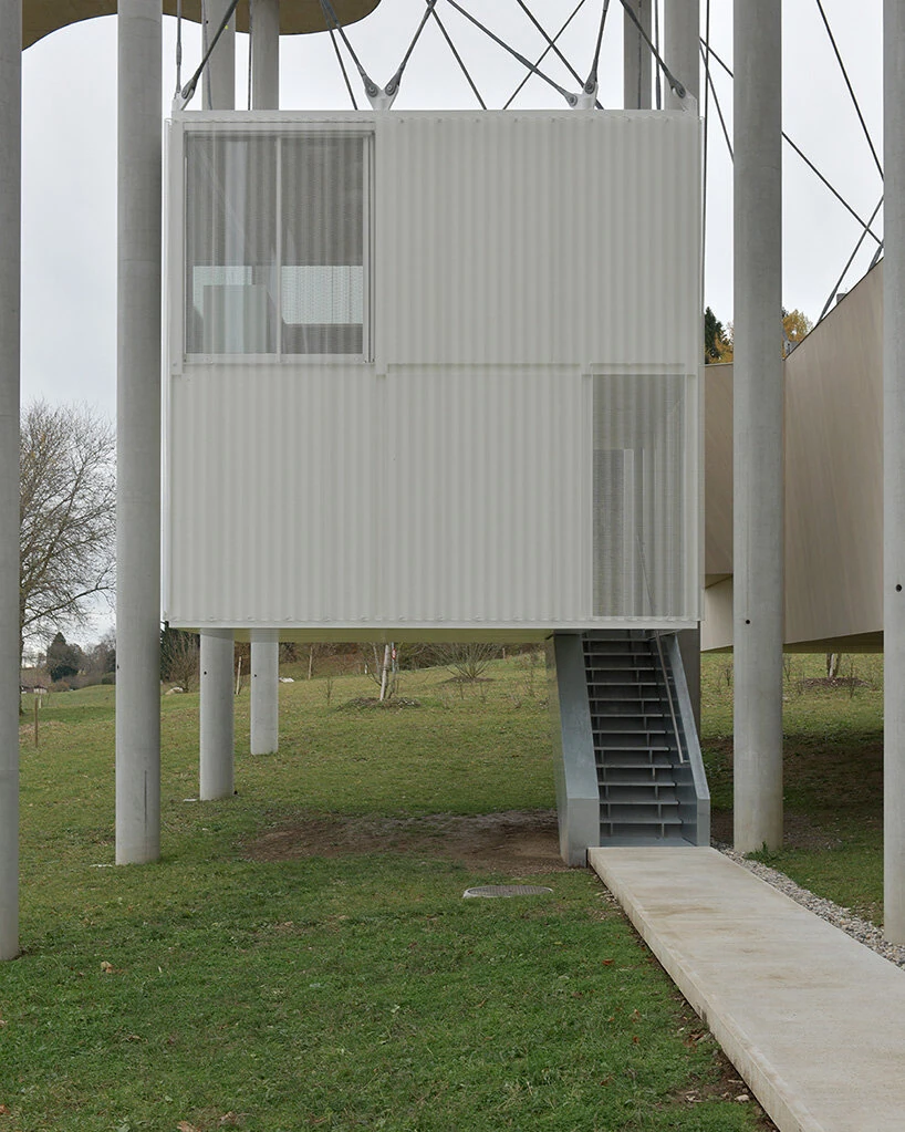 Writers' Cabins at the Jan Michalski Foundation by Kengo Kuma, ELEMENTAL, MK27 and Others, photographed by Leo Fabrizio, Tonatiuh Ambrosetti — atelier bonnet's white cabin, wrapped on four sides in a corrugated skin and translucent mesh screen, stands…