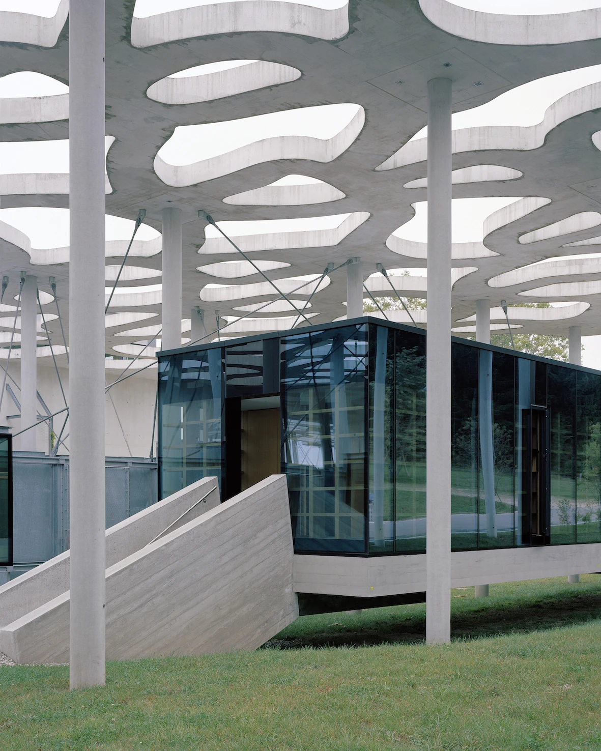 Writers' Cabins at the Jan Michalski Foundation by Kengo Kuma, ELEMENTAL, MK27 and Others, photographed by Leo Fabrizio, Tonatiuh Ambrosetti — ELEMENTAL's tinted-glass cabin reflects the surrounding trees, hung from the perforated canopy on its concrete…
